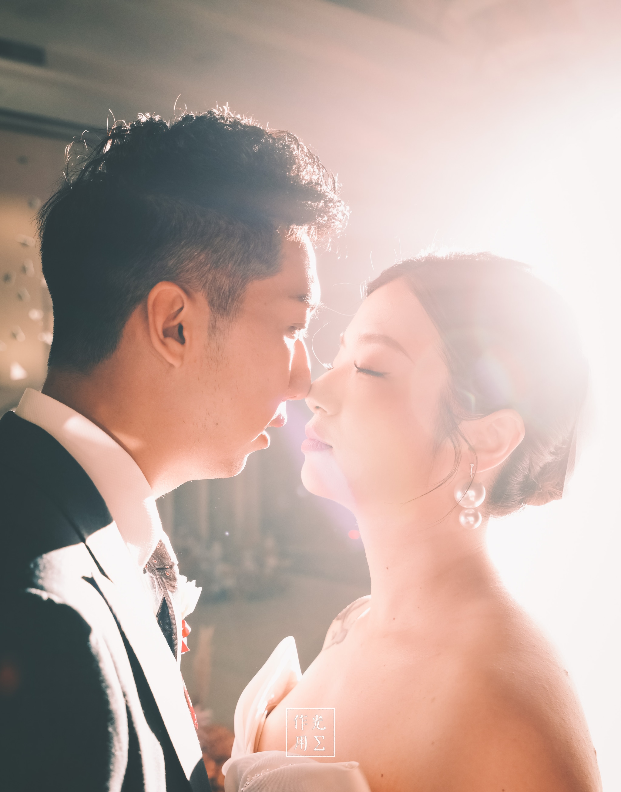 Bathed in a halo of molten light, the bride and groom hover at the edge of a kiss, their profiles melting into a soft-focus glow. His sharp tuxedo lapel and her pearl-trimmed earrings catch stray beams, turning every detail into glinting highlights. Ethereal haze and gentle lens flare fuse the moment into a dreamlike reverie, where time seems to hold its breath