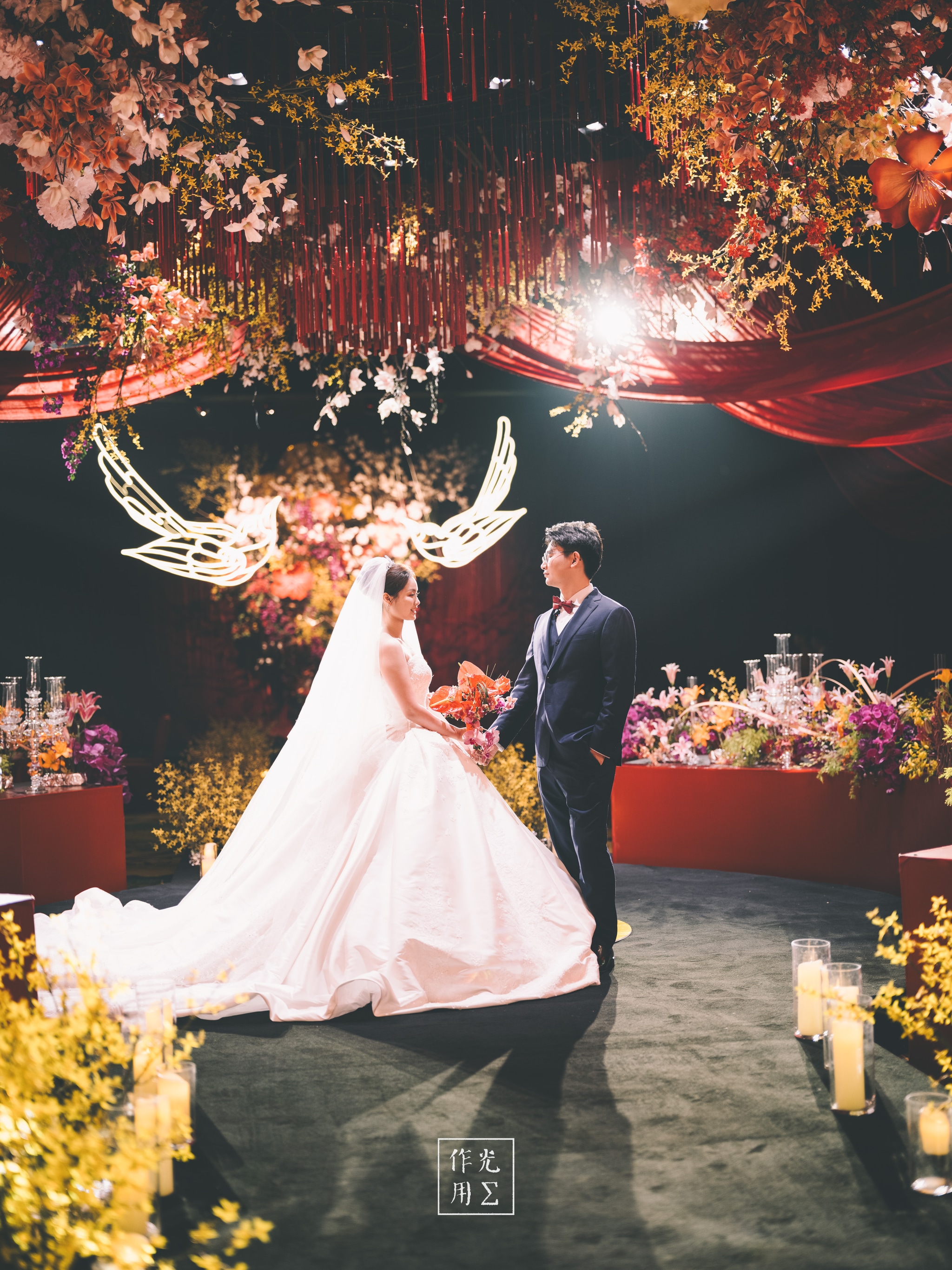 Bathed in a molten amber glow, the couple stands beneath a suspended canopy of blossoms and trailing vines, as if rooted in a secret garden A sculptural antler-like arch, backlit to halo them in soft radiance, frames the bride’s voluminous gown and her cathedral-length veil Flickering candles line the aisle’s edge, their golden light dancing across dark lacquered floors and gilded floral arrangements Rich burgundy drapery and clusters of roses overhead deepen the scene’s warmth, while delicate petals seem to float in midair Every detail—from the bride’s ivory bouquet to the groom’s tailored tuxedo—feels charged with intimate, cinematic romance