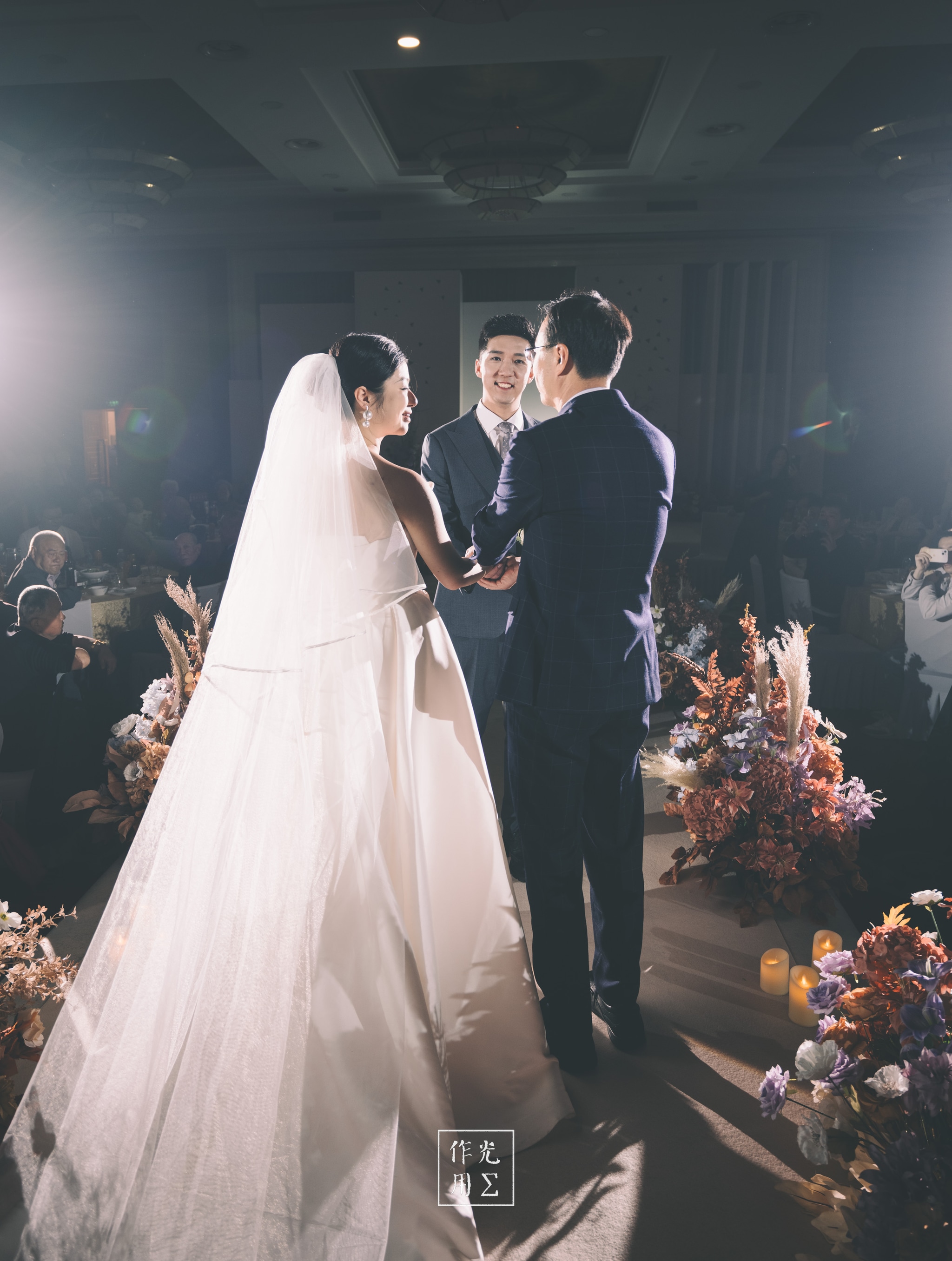 Soft beams of light slice through the dim hall, illuminating the bride’s lace-edged veil as it drapes around her and brushes the aisle. Warm amber spotlights halo the couple and their smiling officiant, casting long, gentle shadows across polished wood floors. Clusters of rust, blush, and ivory blooms flank the altar, their delicate petals catching stray glimmers of light. Gentle haze lingers in the air, giving every moment a dreamlike glow, while seated guests fade into the soft periphery—eyes fixed on the pair exchanging vows beneath that ethereal radiance