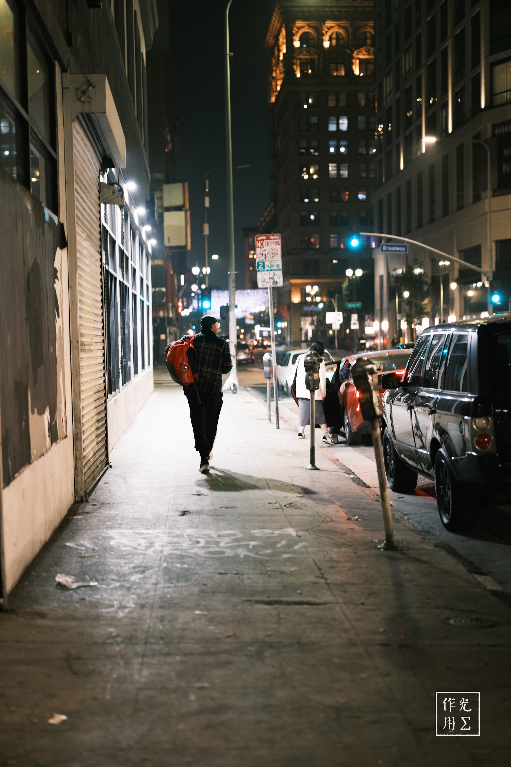 Under the amber glow of streetlights, a solitary figure strides down a damp sidewalk, a scarlet backpack slung over one shoulder. Towering office towers loom overhead, their windows glinting like watchful eyes in the midnight haze. Brake lights along the curb cast ruby reflections on rain-spotted pavement, while distant traffic hums through the quiet. Each echoing footstep cuts through the urban stillness, evoking a blend of melancholy and resolve unique to city nights