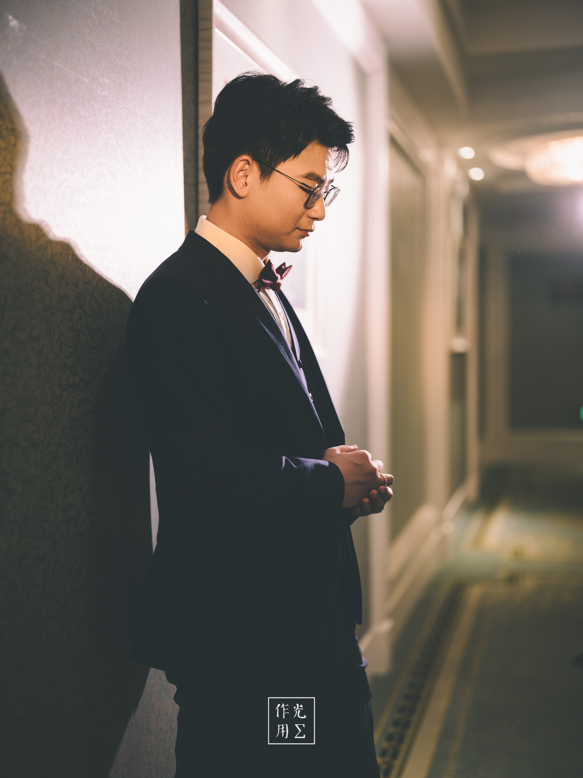 Soft amber light outlines a young man in a tailored black suit and burgundy bow tie, his silhouette pressed against the hotel corridor’s cool gray wall. A shallow focus blurs the distant lamps and patterned carpet into soft bokeh, amplifying his quiet poise as he scrolls on his phone. The muted palette—warm highlights melting into dusky shadows—imbues the scene with contemplative stillness, each detail sharpening the contrast between his refined attire and the hushed, endless corridor