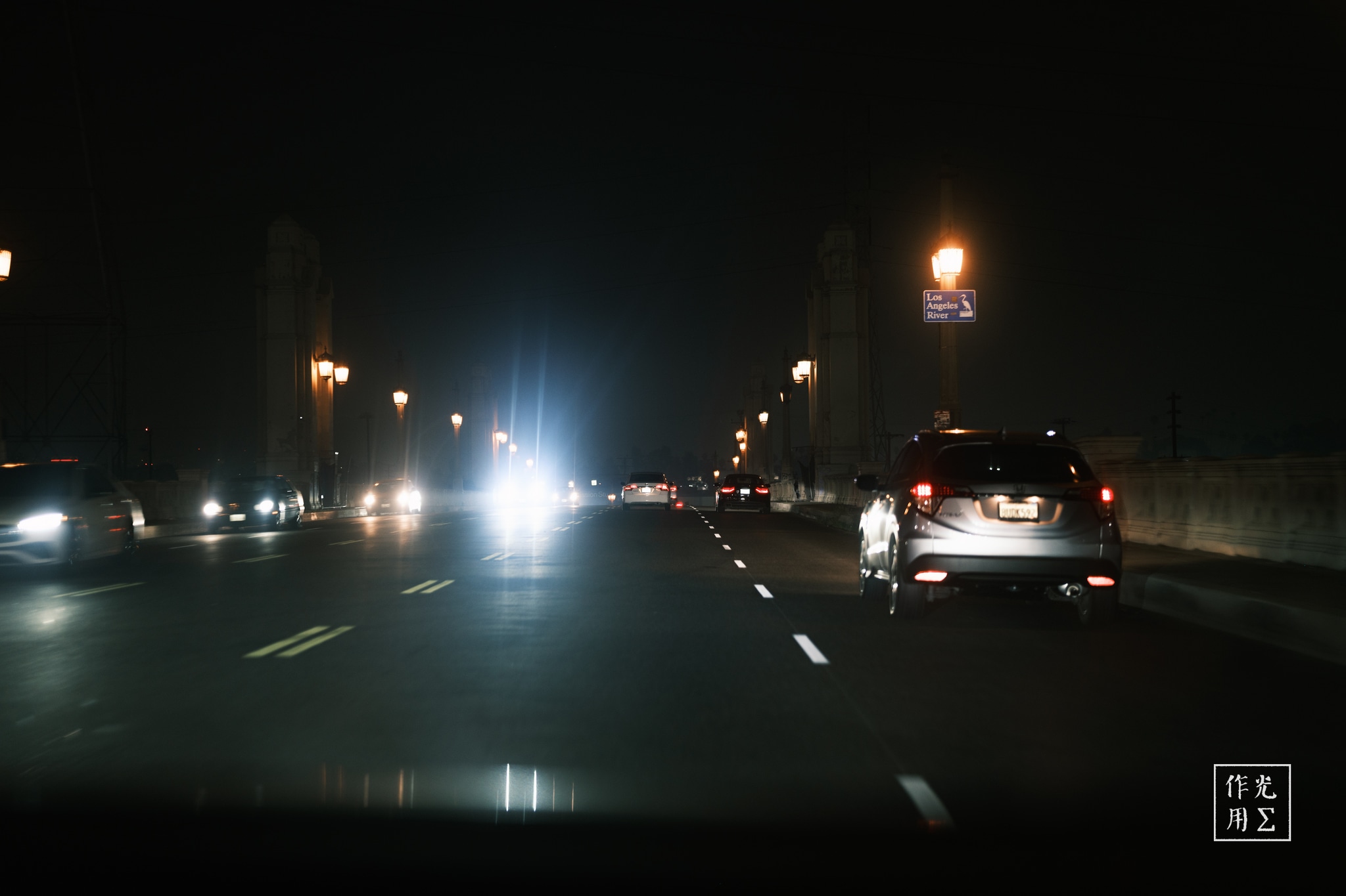 Luminous headlights carve through the ink-black asphalt, their silver beams fracturing into glinting ribbons that dance across the wet road. Lantern-warm streetlamps punctuate the deep gloom, their soft halos warping against a silent brick wall. A lone sedan idles at the curb, its sleek silhouette frozen in the hush between passing cars—a taut moment of urban solitude, alive only in light and shadow