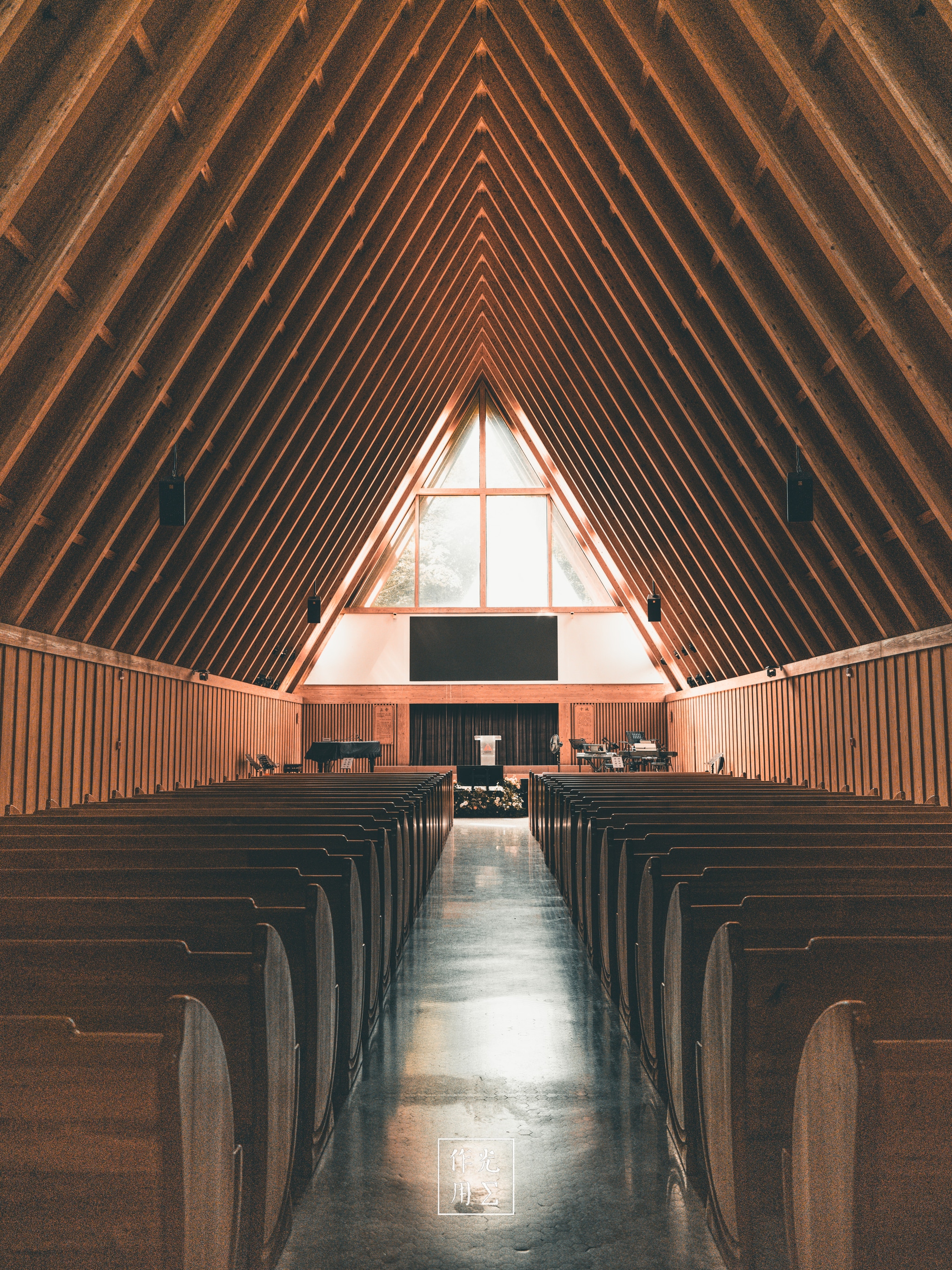 Sunlight streams through the soaring triangular window, casting a warm amber glow along the polished aisle. Slender wooden beams rise in perfect symmetry, converging overhead like silent sentinels. Rows of simple, curved pews flank the center walkway, their smooth grain accentuated by the soft, diffused light. At the far end, a modest altar bathed in gentle radiance anchors the space, lending an air of hushed reverence. The hush between the rafters and the golden hue creates a calm, almost meditative atmosphere—an architectural hymn in wood and light