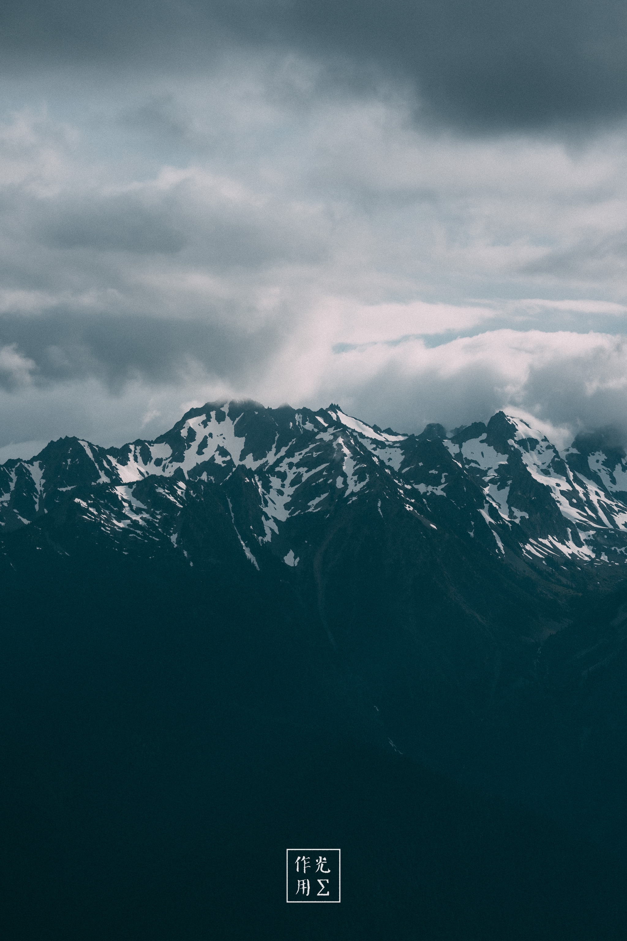 Jagged peaks thrust skyward, their dark flanks streaked with filigrees of snow that gleam like molten silver against bruised clouds. A heavy, brooding sky churns above, the low-hanging storm clouds parting in places to spill pale shafts of light onto the craggy ridges below. Mist clings to the mountain’s shoulders, softening the sharp silhouettes and lending a sense of shifting scale—as if the peaks might vanish back into the clouds at any moment. The overall palette of slate blues and steely grays deepens the mood, turning the scene into a raw, elemental tableau of rock, ice, and brewing tempest
