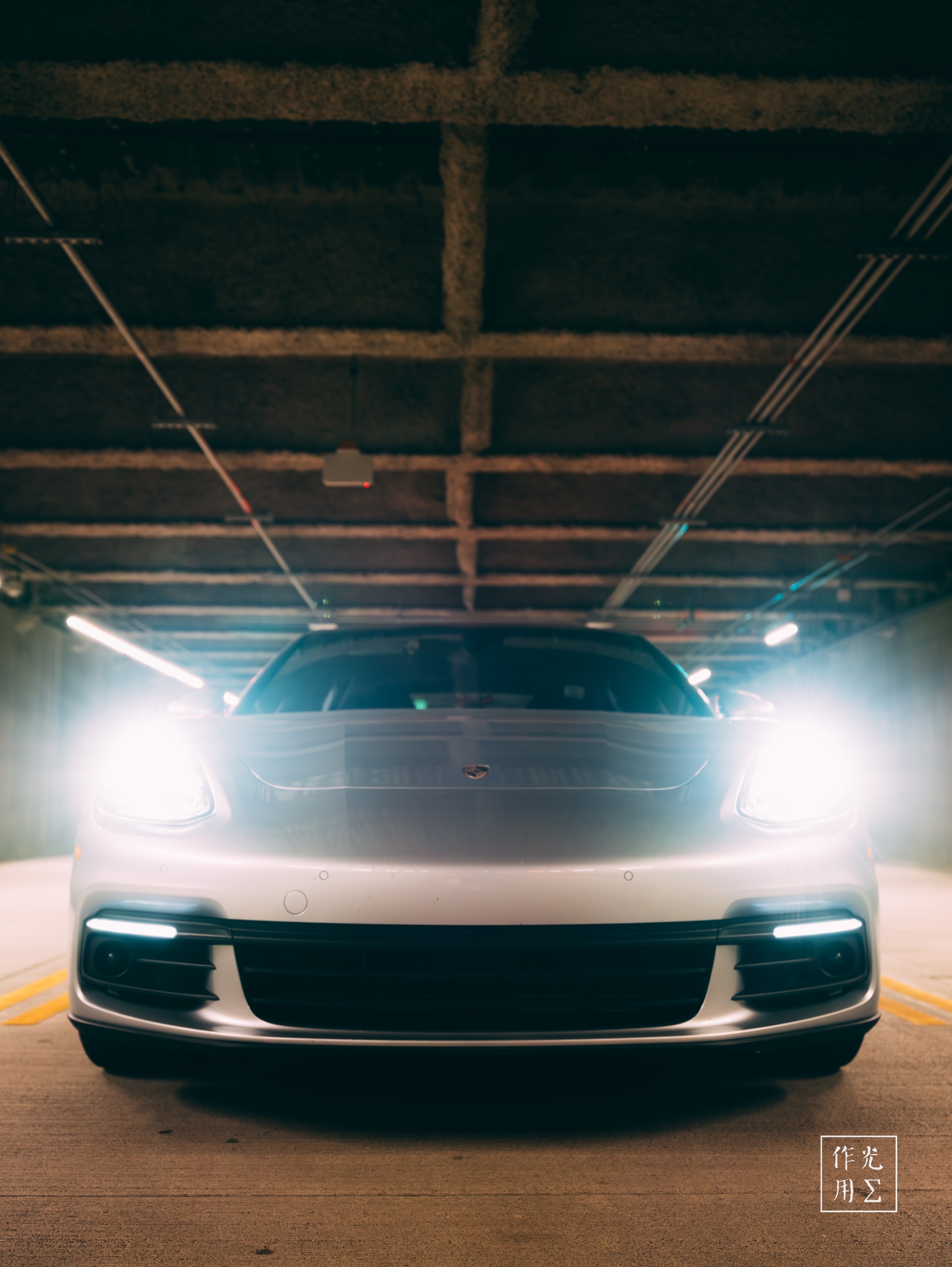 Piercing LED beams slice through the garage’s smoky half-light, casting the low-slung coupe in sharp, spectral highlights. Concrete girders overhead form a grid-iron vault that contrasts with the car’s mirror-smooth curves. A near-perfect symmetry and the raw, industrial setting infuse the scene with cinematic tension, as if this electric stalker is moments from springing to life
