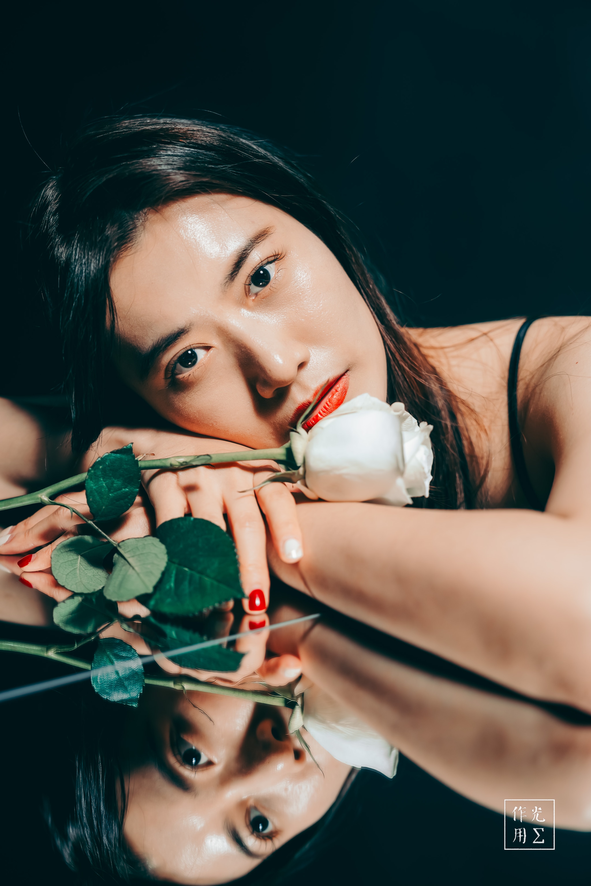 Soft lamplight carves gentle shadows across her porcelain skin as her fingertips brush the petals of an ivory rose. Her mirrored gaze in the glass plane doubles the intimacy, the reflection echoing unspoken longing. A ribbon of crimson on her lips punctuates the near-monochrome hush, infusing the scene with quiet drama and fragile elegance