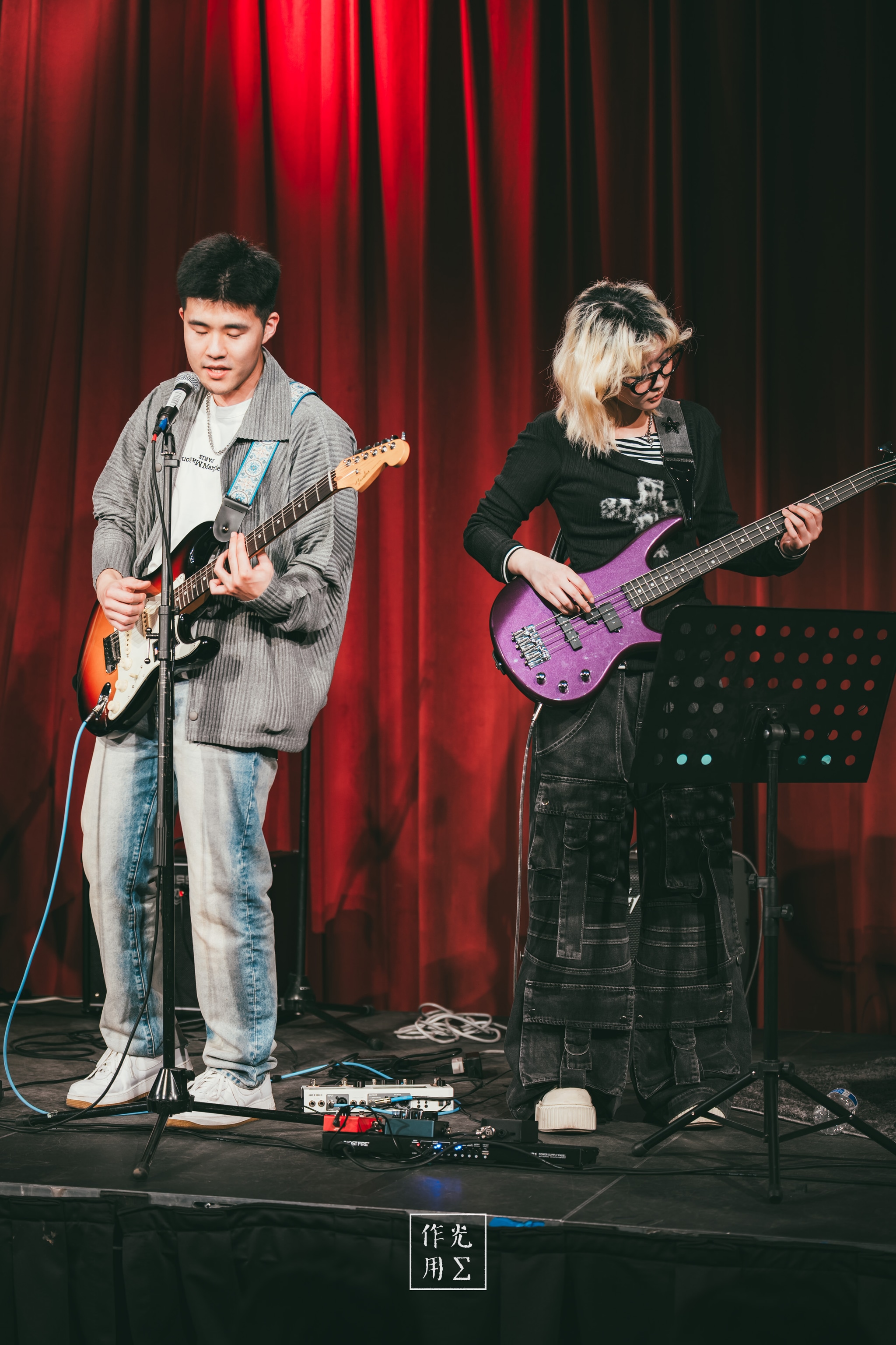 Bathed in moody scarlet light, a young guitarist leans into his microphone, fingers dancing across sunburst frets while his bandmate anchors the groove with a glossy purple bass. Velvet red curtains ripple behind them, framing the duo in an intimate spotlight. A tangle of pedal effects and cables at their feet hints at raw experimentation, and their casual streetwear—oversized jackets and chunky sneakers—contrasts with the stage’s theatrical backdrop. The tension between earnest concentration on the singer’s face and the bassist’s cool poise gives the scene an undercurrent of youthful defiance