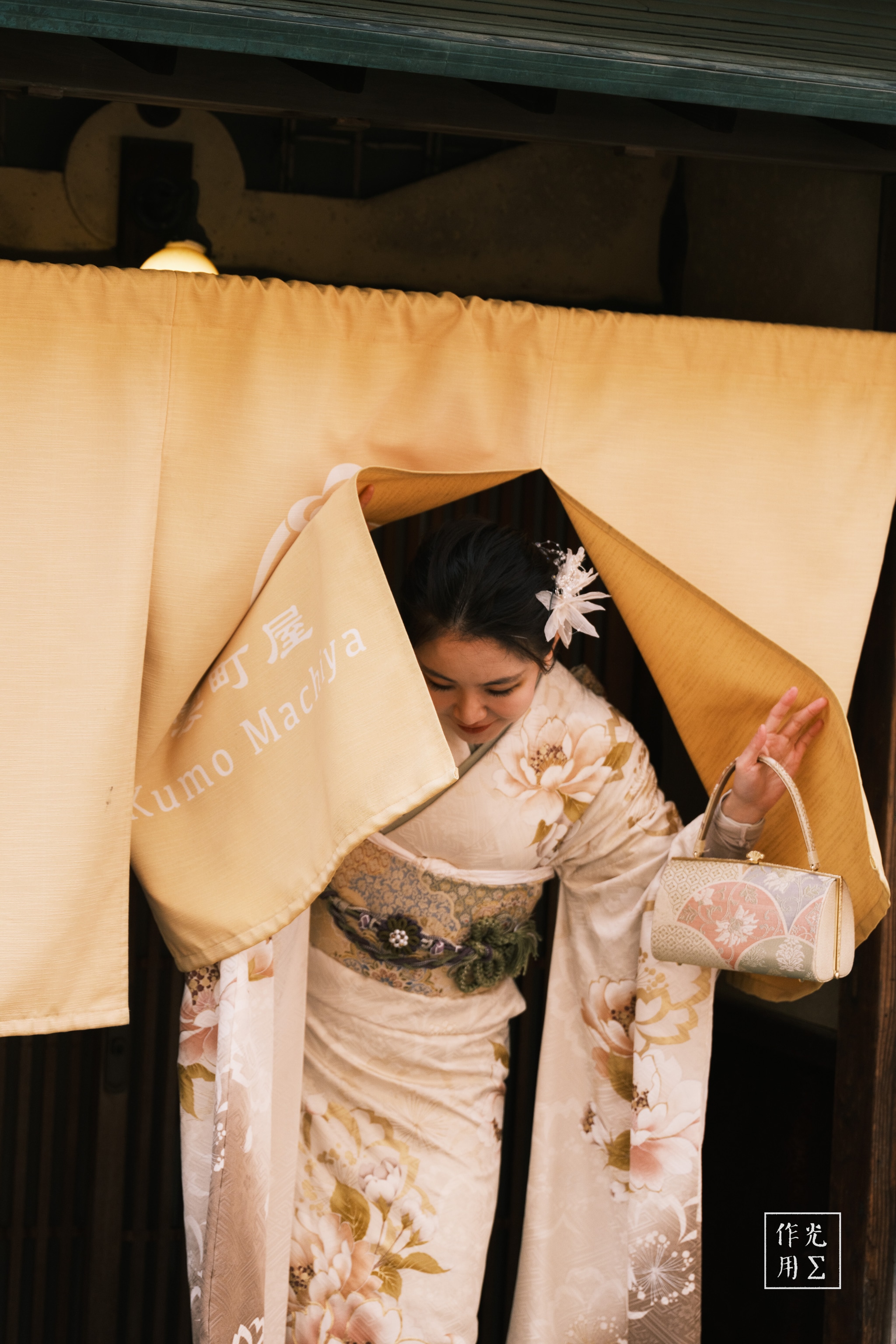 Sliding apart the ochre noren, she emerges swathed in an ivory kimono patterned with pale chrysanthemums, the silk’s soft sheen catching every flicker of the lantern light. Her obi’s intricate weave cinches the robes at the waist, while a single white blossom graces her dark hair, lending a whisper of spring to the warm, hushed entryway. The amber glow and tender tension in her downward gaze conjure a moment suspended between tradition and quiet anticipation