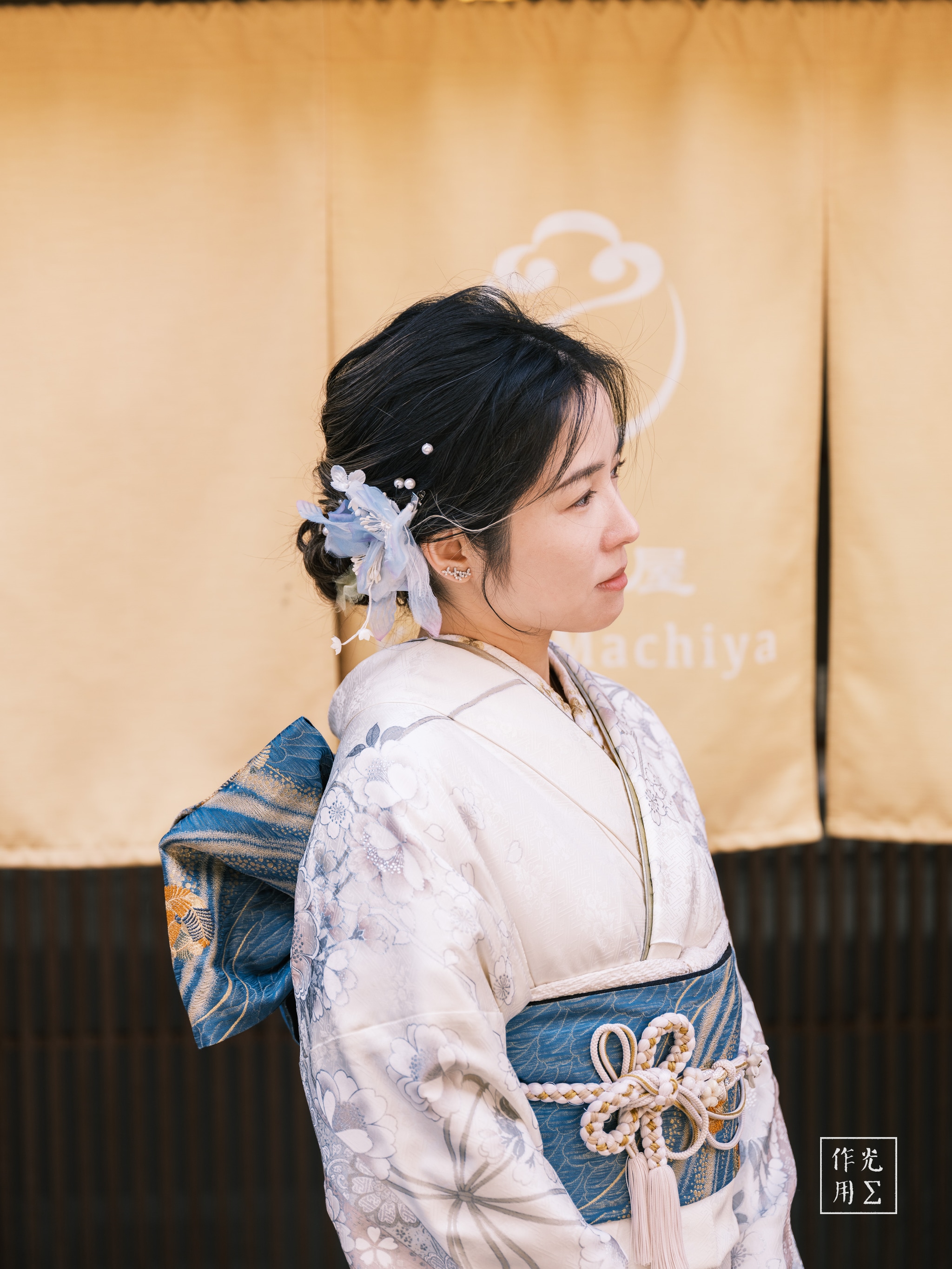 Bathed in soft, golden light, her ivory kimono shimmers with silver-threaded florals that ripple across the fabric like moonlit water. A deep indigo obi, cinched with an intricate mizuhiki knot, anchors the ensemble, while delicate silk blossoms and gently cascading ribbons tuck into her dark hair. Her sideways glance, serene and introspective, brings out the quiet grace of the moment. Behind her, a muted ochre curtain embossed with a stylized cloud motif frames the scene, lending warmth and a subtle air of timeless tradition