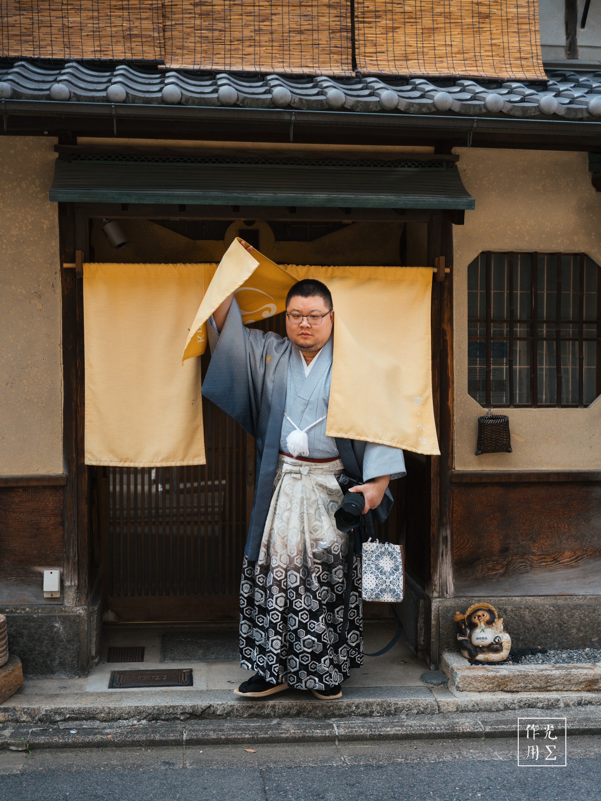 Stepping through a cream-colored noren, a figure in a pale-blue kimono and patterned hakama lifts a woven kasa hat, sunlight catching his glasses and revealing a calm, confident gaze. Behind him, weathered wooden beams, earthen plaster walls and a latticed window frame his silhouette, while a small ceramic tanuki statue crouches by the doorstep. The warm glow of afternoon light softens the scene, blending time-worn architecture with the quiet dignity of traditional dress