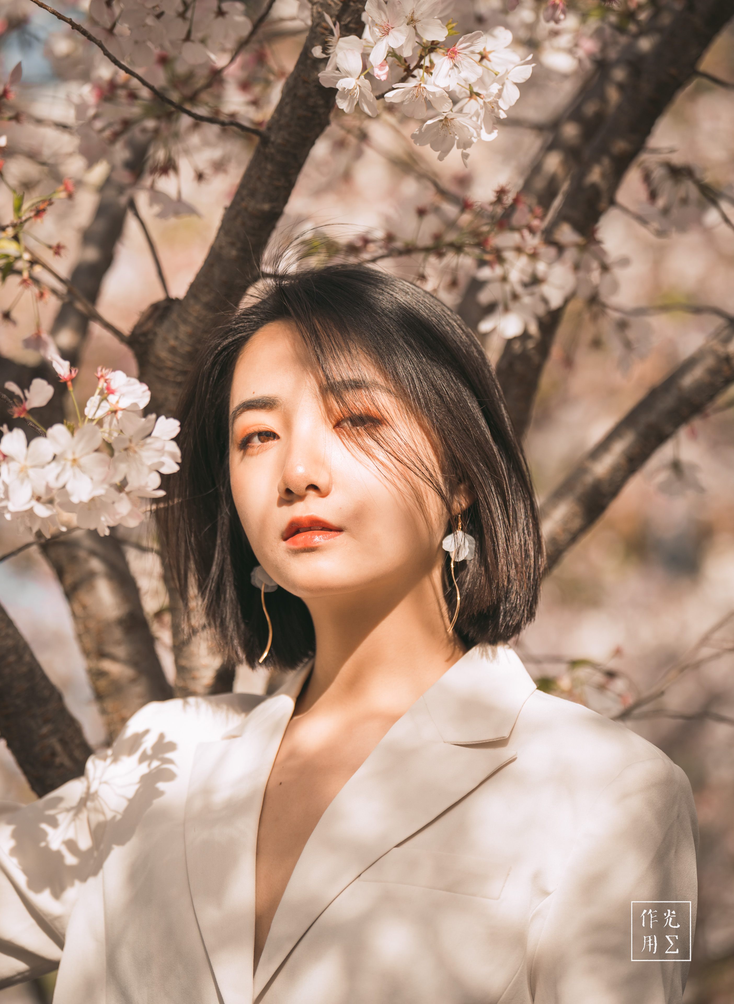 Sunlight dances through pale-pink blossoms, casting a warm glow across her poised silhouette. Soft shadows ripple over the ivory blazer, where sleek lines of tailored fabric meet the gentle curves of drifting petals. Her calm, introspective gaze and dangling geometric earrings anchor the scene in quiet confidence, while blurred branches above create an ethereal canopy of spring’s delicate bloom