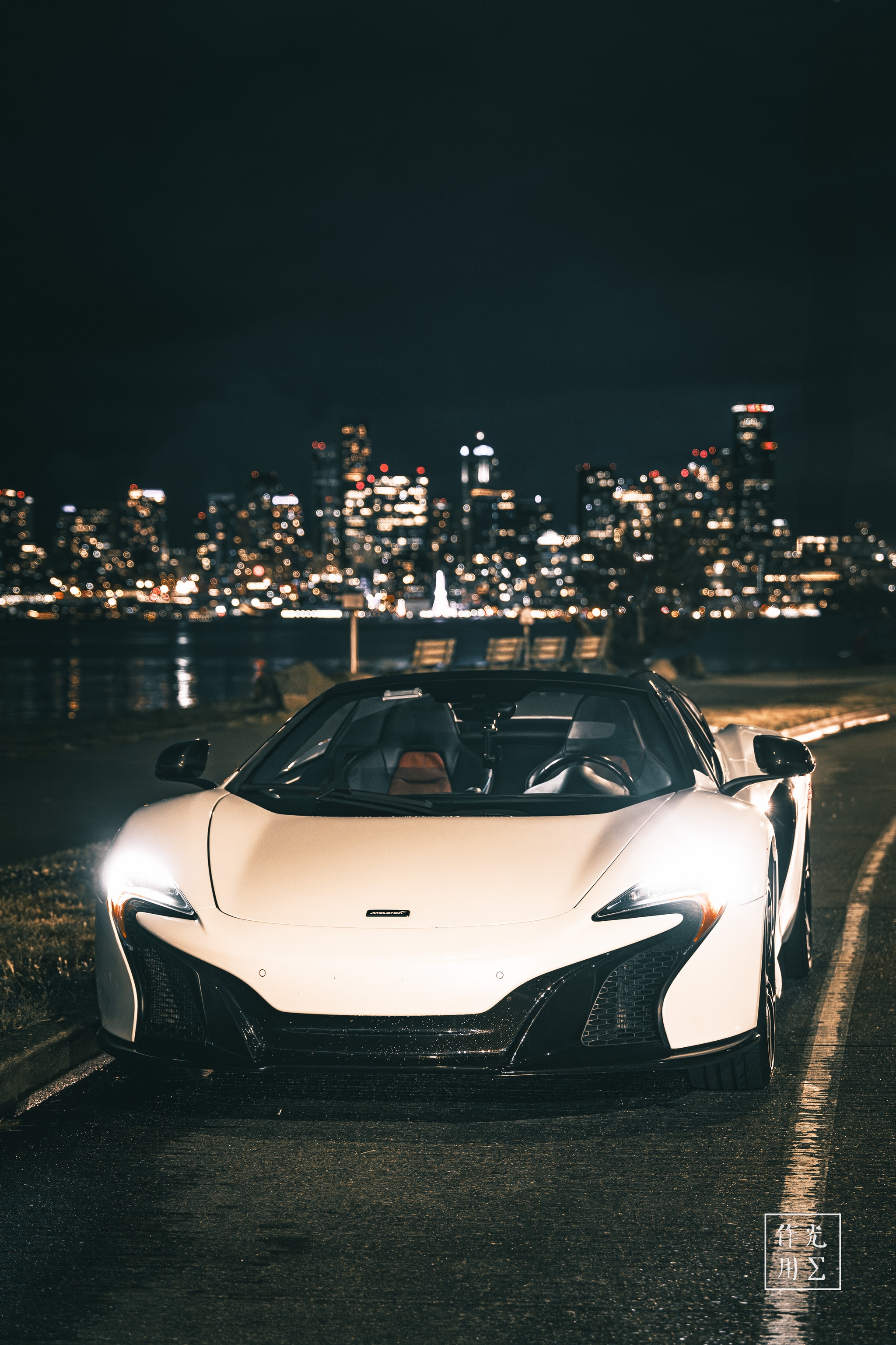 Glancing steel-white curves hug the pavement as twin LED “eyes” slice through the midnight haze. Gleaming carbon accents catch stray city sparks, while ripples of harbor light dance across the hood. Behind the poised McLaren, a constellation of skyscraper windows shimmers on dark water, lending the scene an electric tension—sleek power waiting in silent communion with the urban glow