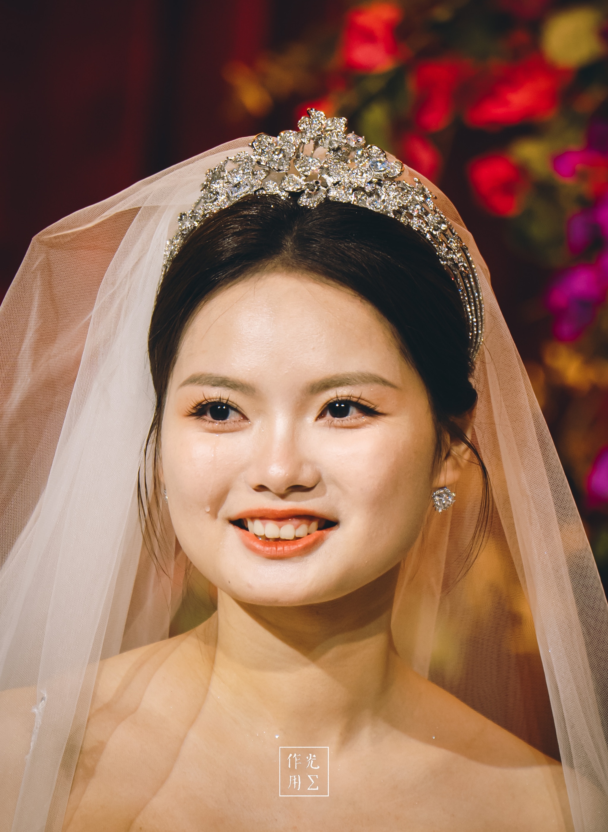 Soft golden light dances across her flawless skin as a crystal-encrusted tiara crowns her dark hair, each gem catching the glow Silk veil drapes in whisper-thin layers, framing her serene smile and the gentle curve of her collarbone Behind her, deep scarlet blooms blur into painterly strokes, their rich petals echoing the warmth in her eyes The scene pulses with quiet joy, every shimmer and hue lending an air of radiant celebration