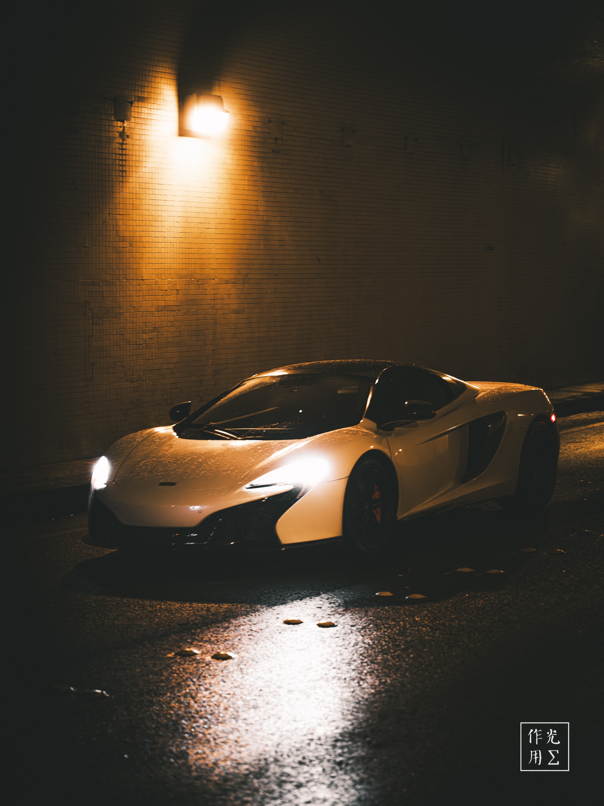 Glinting under a lone amber bulb, the low-slung supercar’s sculpted fenders and sweeping curves emerge from the tunnel’s inky gloom. Droplets on its glossy bonnet catch faint reflections from the slick pavement, while the surrounding concrete walls absorb and diffuse the warm glow into soft gradients of shadow. The scene feels electric yet hushed—an intimate, almost cinematic pause where power and stillness coalesce in a secret urban sanctuary