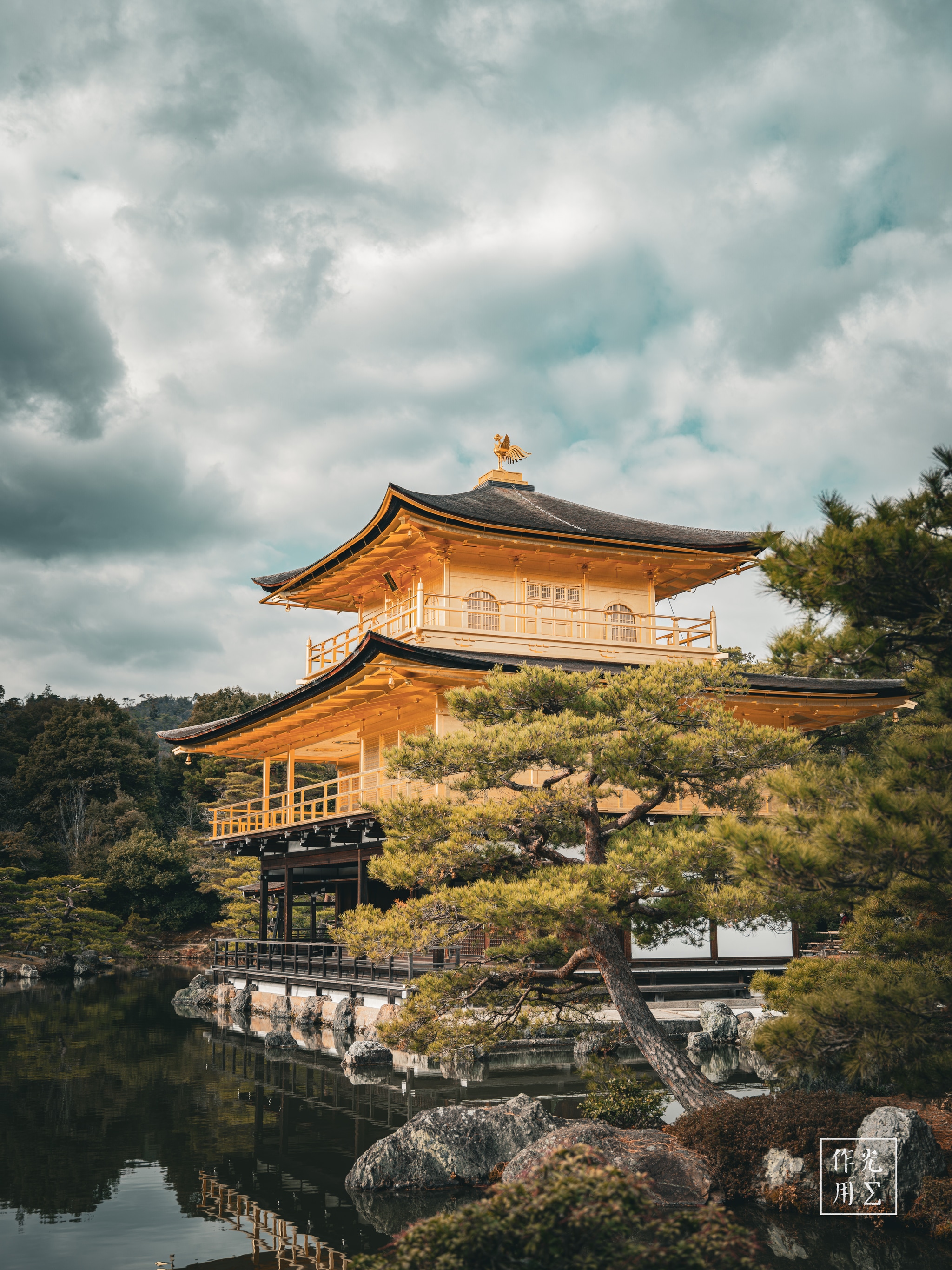 Gleaming golden tiers hover above a glass-smooth pond, their lacquered eaves catching the muted glow of an overcast sky. Gnarled pine branches frame the pavilion’s ornate silhouette, while ripples of emerald water mirror the temple’s gilded warmth against the brooding charcoal clouds. Moss-clad rocks and slender wooden walkways trace a serene path around the perimeter, inviting quiet reflection in the hush before an impending rain