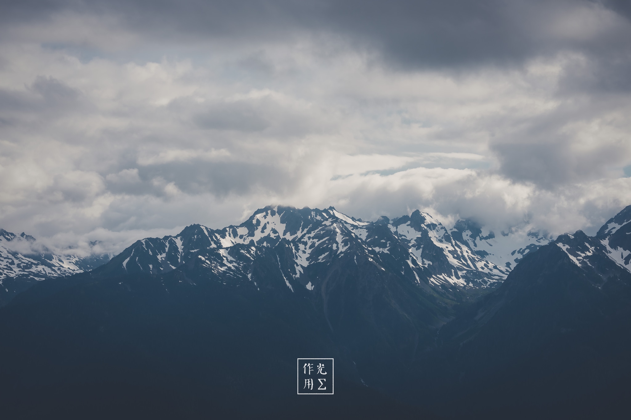 Jagged peaks slice through low-hanging clouds, their snow-spattered ridges gleaming like fractured mirrors against a slate-gray sky. Rolling banks of mist coil around each summit, lending an otherworldly hush that heightens the sense of raw, untamed grandeur. A brooding tension hangs in the air, as if the mountains themselves are holding their breath before an approaching storm