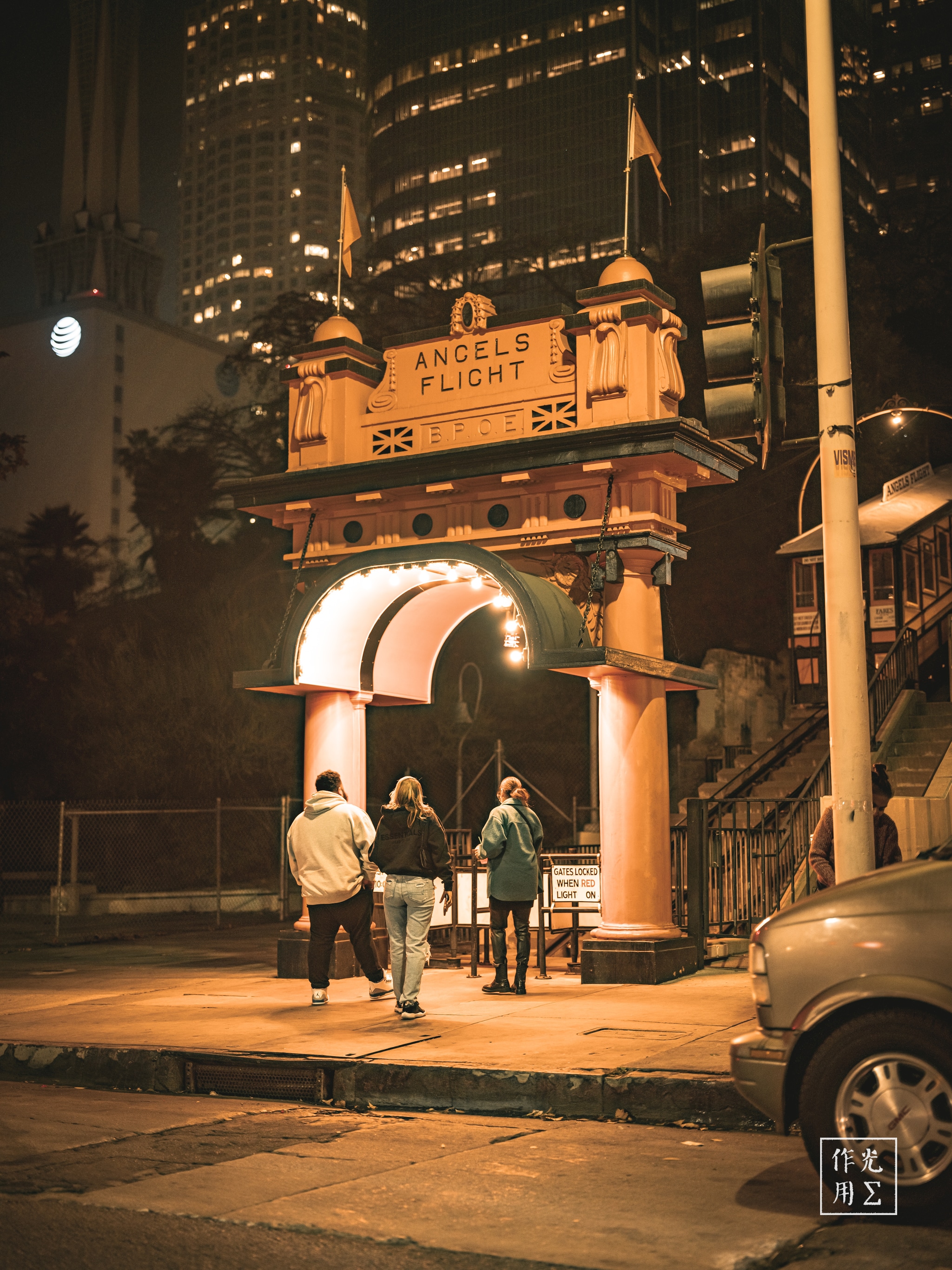 Bathed in honeyed streetlight, the century-old Angels Flight archway gleams against a backdrop of glass-and-steel skyscrapers. Soft shadows dance across its red-and-gold façade, where the peeling paint and ornate finials whisper of countless ascents and descents. Three figures pause beneath the arched entry—jackets rustling in a gentle breeze—as they gaze upward, caught between the nostalgia of a bygone era and the humming pulse of the modern city. The warm glow, muted hum of traffic, and the promise of a brief, vertiginous ride combine into a cinematic moment suspended in time
