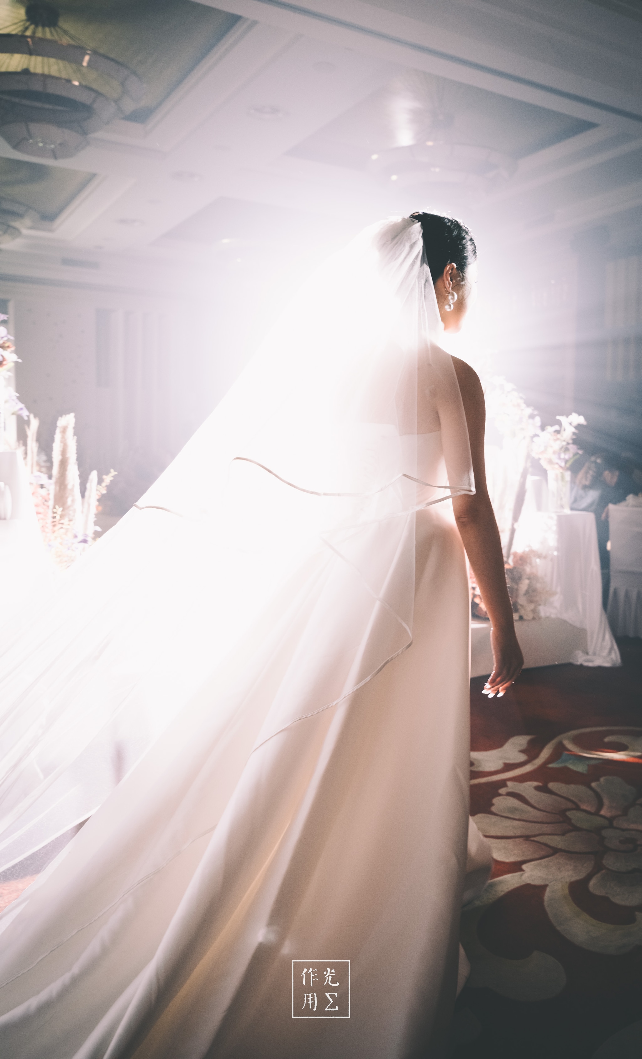 Ethereal light floods the grand ballroom, casting her flowing veil into a halo of radiant white. The ivory gown drapes in sculptural folds across the patterned carpet while crystal chandeliers glimmer overhead. An expectant hush hums through the air as daydream and reality blur under the luminous glow