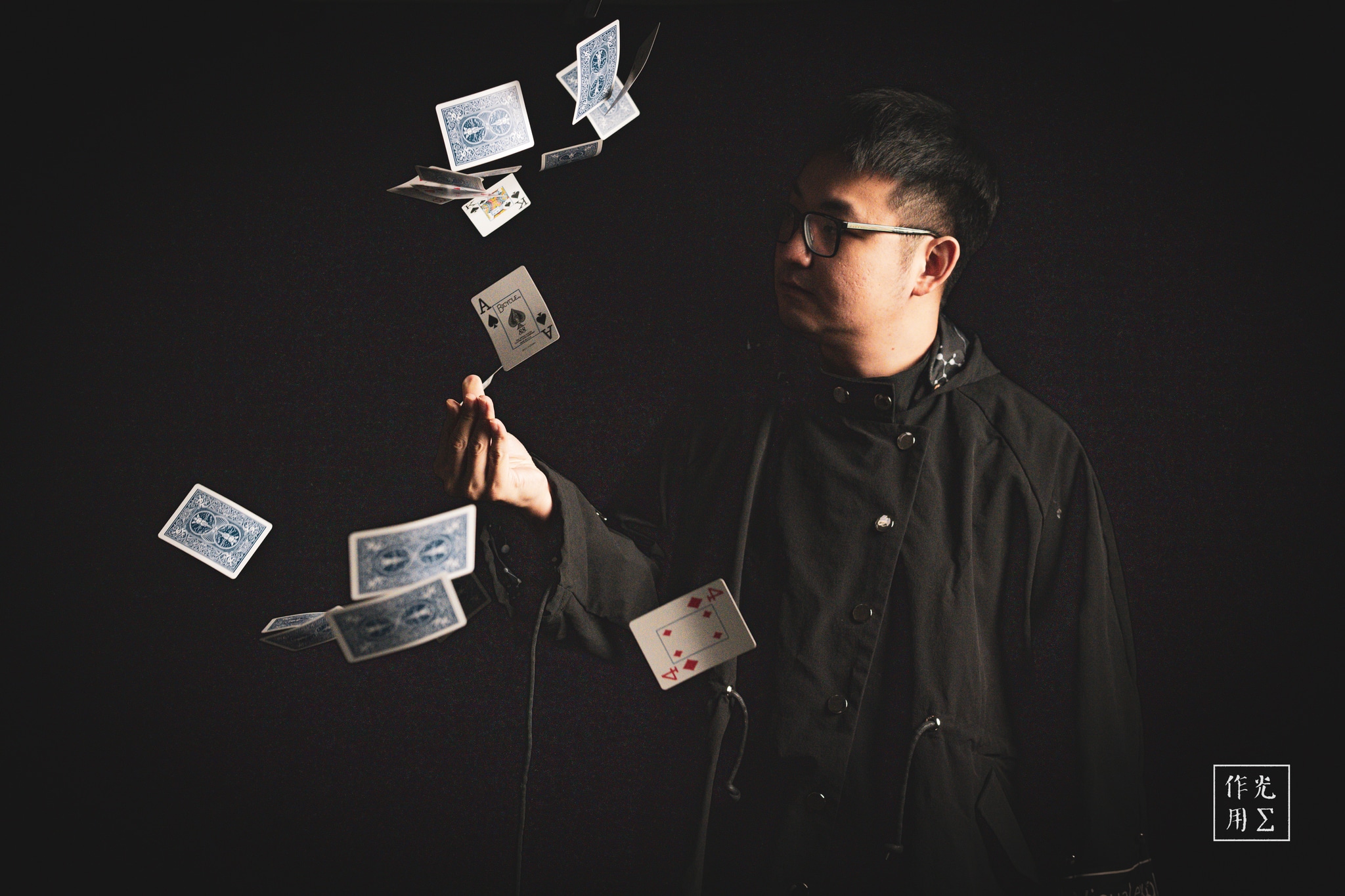 Commanding the darkness with a single, outstretched wand, a young magician conjures a graceful spiral of playing cards that seem to float in mid-air. Sharp side lighting carves his focused profile and the crisp edges of each card against the inky black backdrop. His charcoal shirt and round glasses lend an aura of understated poise, while the cards’ white faces and bold pips shimmer like fragments of starlight. The scene pulses with quiet suspense, as if time itself has paused to watch this delicate dance of sleight and shadow