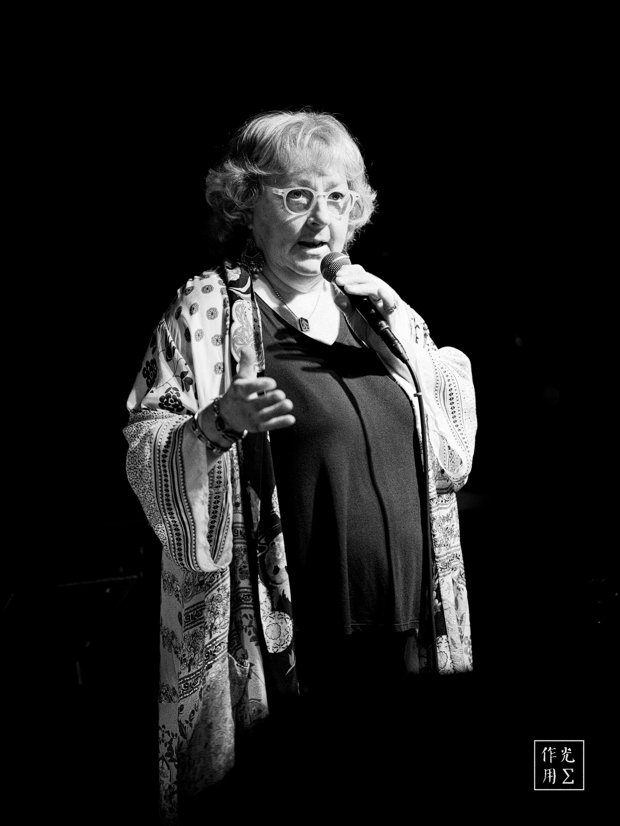 Floodlit against an inky void, a silver-haired storyteller grips the microphone with poised intensity. Wispy curls frame wire-rim glasses that catch the spotlight’s gleam, while a flowing, kimono-style robe patterned in subtle florals drapes around her slender frame. Every gesture—one hand gently raised, the other anchoring the mic—radiates quiet authority. Deep shadows carve dramatic contours across her face, lending the scene an intimate, almost cinematic gravitas