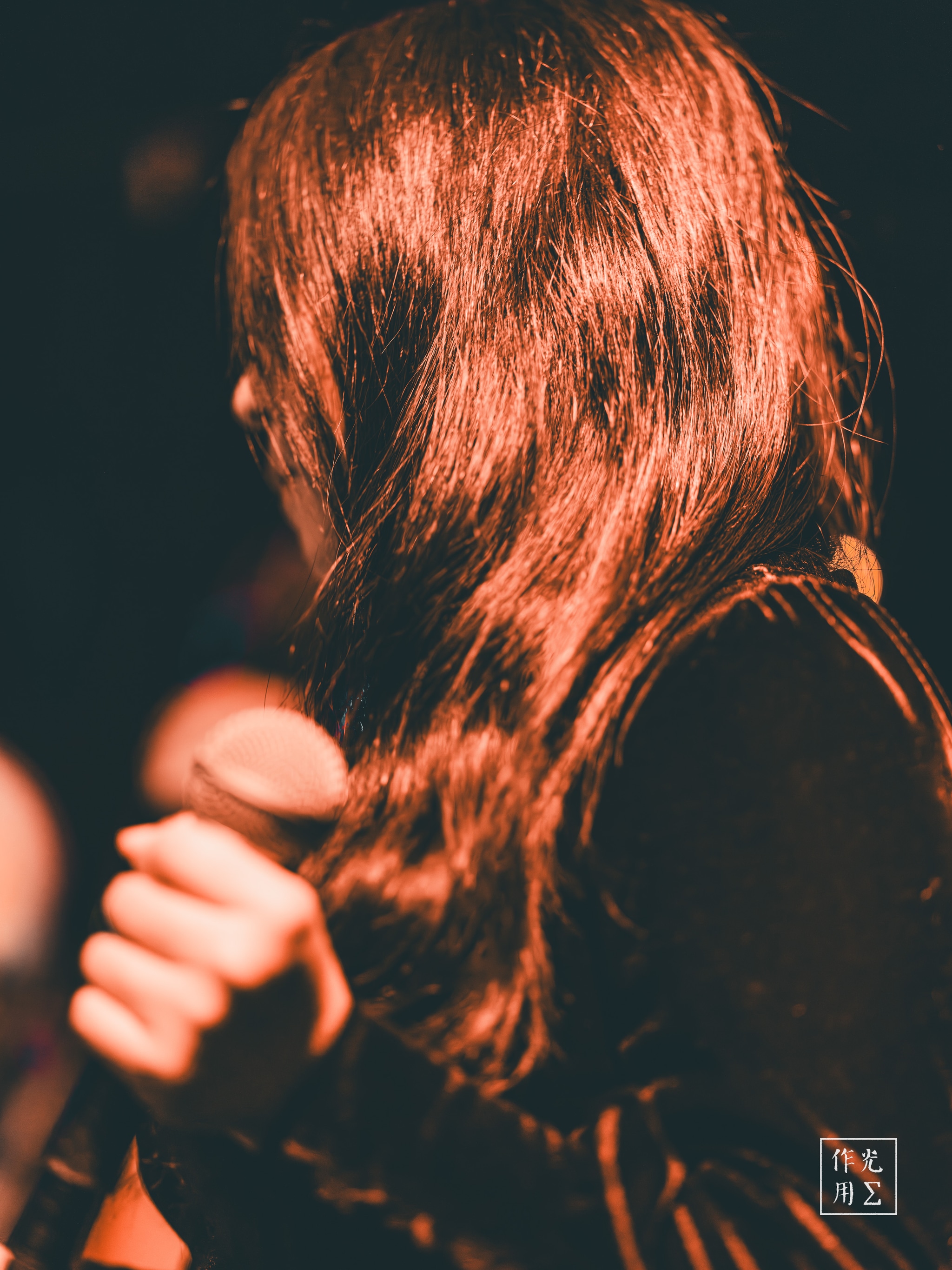 Cascading auburn waves glimmer under a warm spotlight, each strand catching the stage’s copper glow. A slender hand grips a vintage microphone, knuckles white against its polished metal. Velvet sleeves absorb the surrounding darkness, lending depth to the scene’s rich, sensuous palette. Hints of distant bokeh lights swirl behind her, as if the audience’s heartbeat has been distilled into glowing orbs. The air feels thick with anticipation, charged by the promise of a voice about to rise