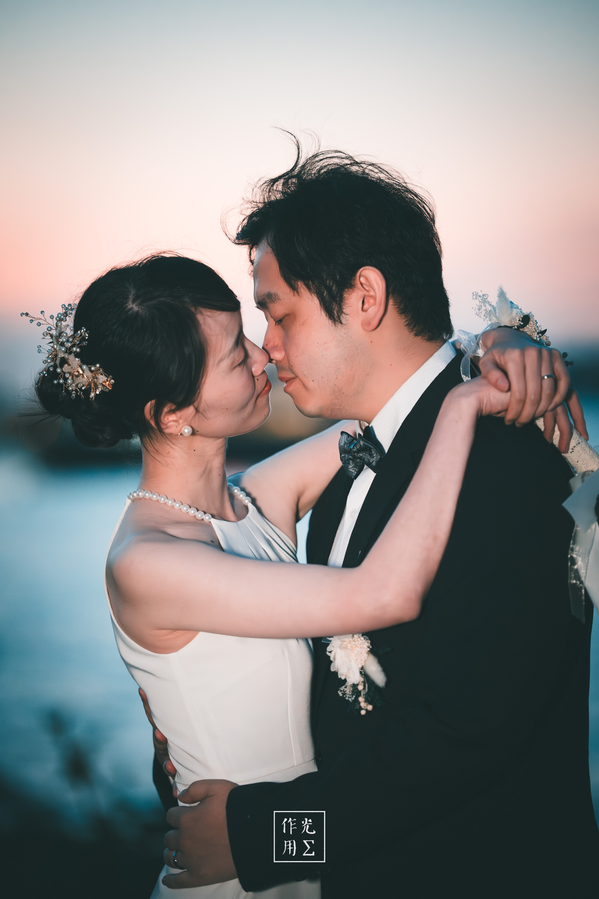 Soft twilight drapes a serene lakeshore as the newlyweds melt into each other’s arms, foreheads nearly touching Her ivory gown catches the blush of sunset while a delicate floral hairpin and pearl necklace glimmer against soft waves His black tuxedo frames her in stark relief, every gentle sway igniting warmth in their quiet, breathless moment