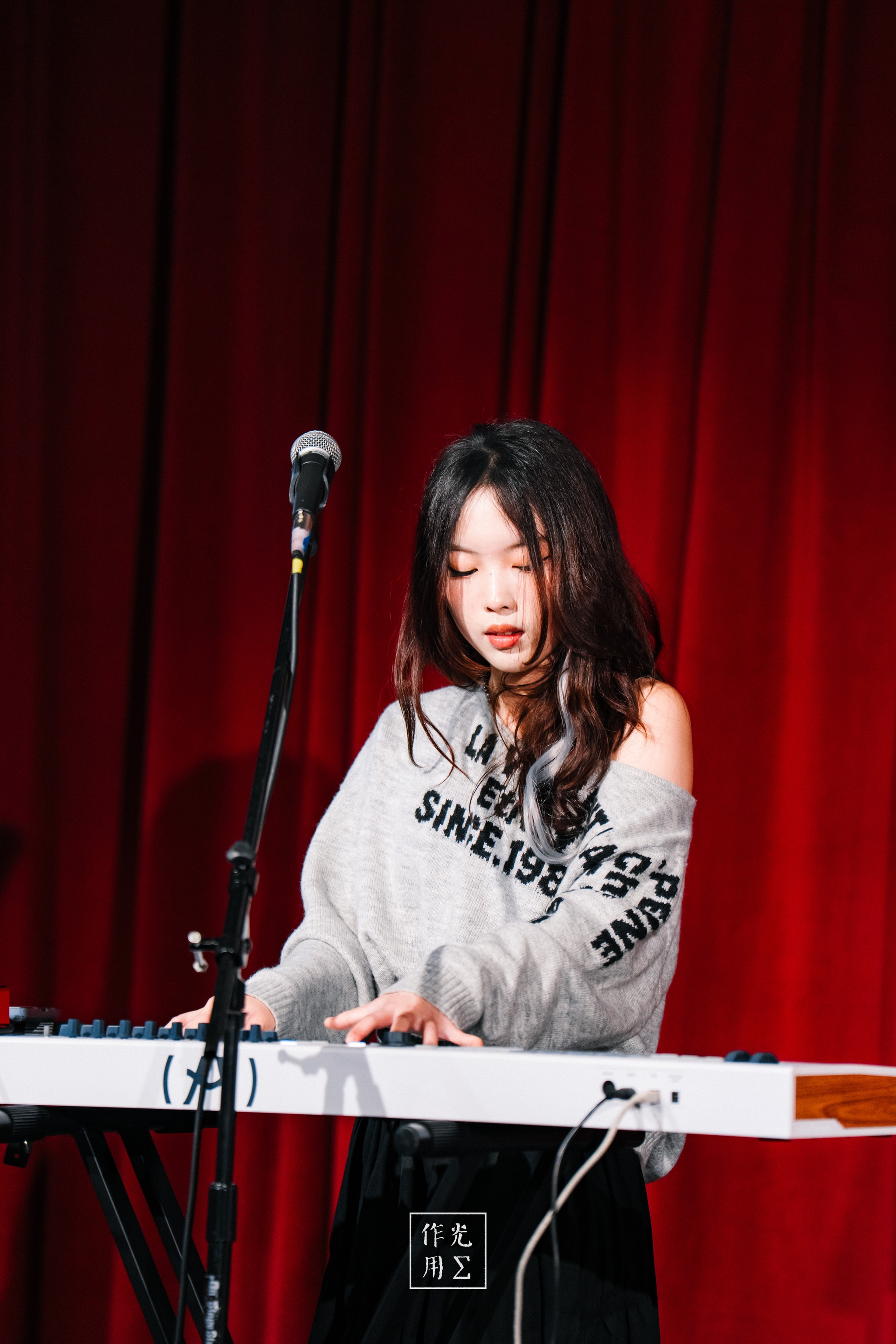 Fingers glide across ivory keys beneath a warm spotlight, each note unfurling into the deep crimson velvet behind her. Loose black waves frame a calm, concentrated expression as the off-shoulder gray sweatshirt—lettering slanted in bold black—hints at casual confidence. A single mic stand waits expectantly to capture her breathy vocals while slender hands coax melodies that rise and fall like whispered secrets. The rich red drape absorbs and reflects the light, giving her silhouette a painterly glow. Subtle shadows cradle the piano’s sleek white surface, and every detail—from the delicate tilt of her chin to the gentle sway of her sweater—feels charged with intimate energy. The scene pulses with a quietly electric tension, as if the air itself listens