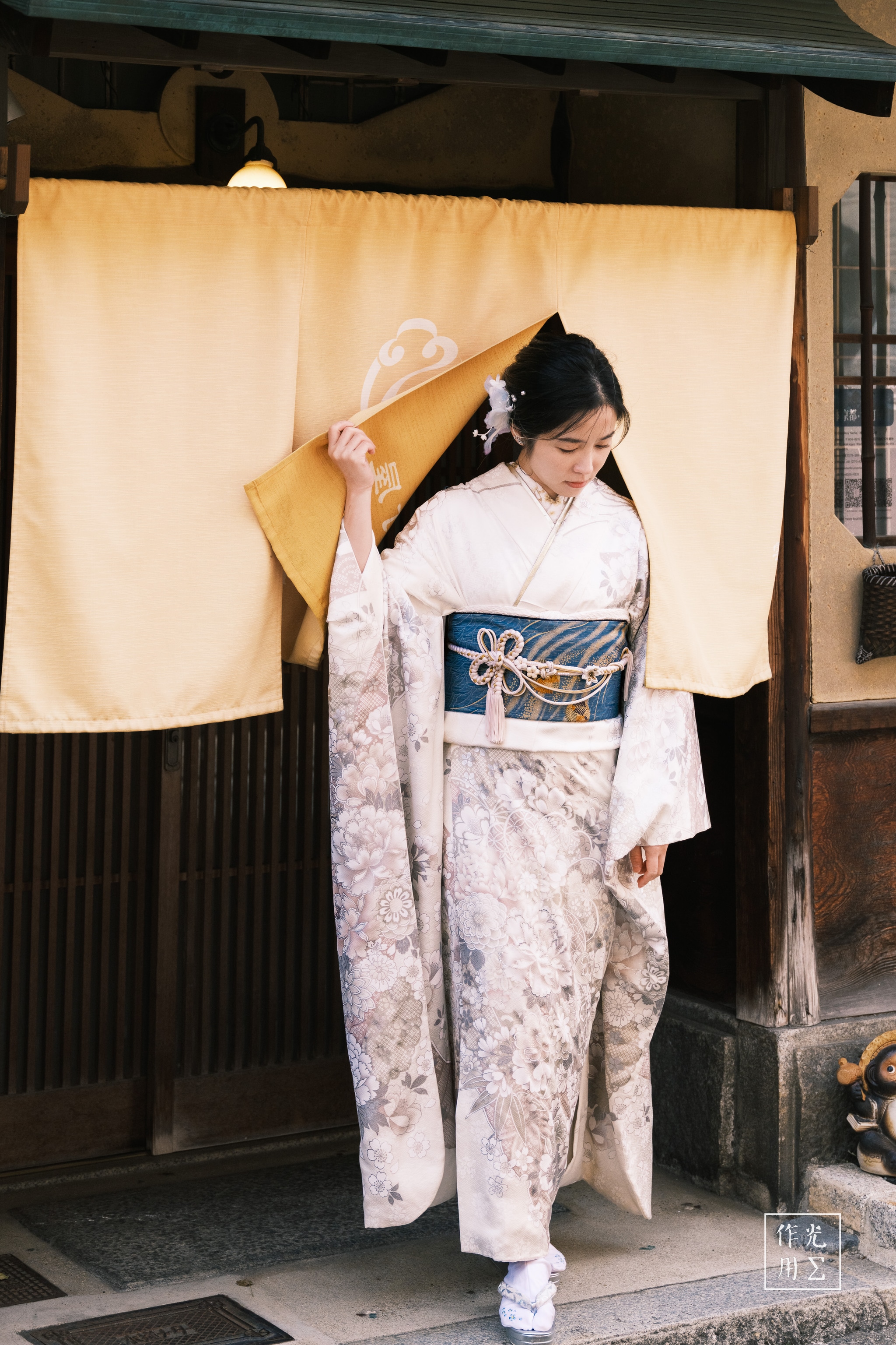 Lifting the soft ochre noren with poised fingers, a young woman in a pale kimono pauses at a weathered wooden threshold. Intricate plum blossoms unfurl across her ivory silk, while a deep indigo obi cinched with coral cords anchors the ensemble in bold relief. Slanted afternoon light filters through latticed windows, casting warm shadows onto the earthen façade and a tiny ceramic fox at her feet. Her lowered gaze and the gentle ripple of fabric conjure a hushed, timeless moment—where tradition, texture, and muted color converge in quiet harmony
