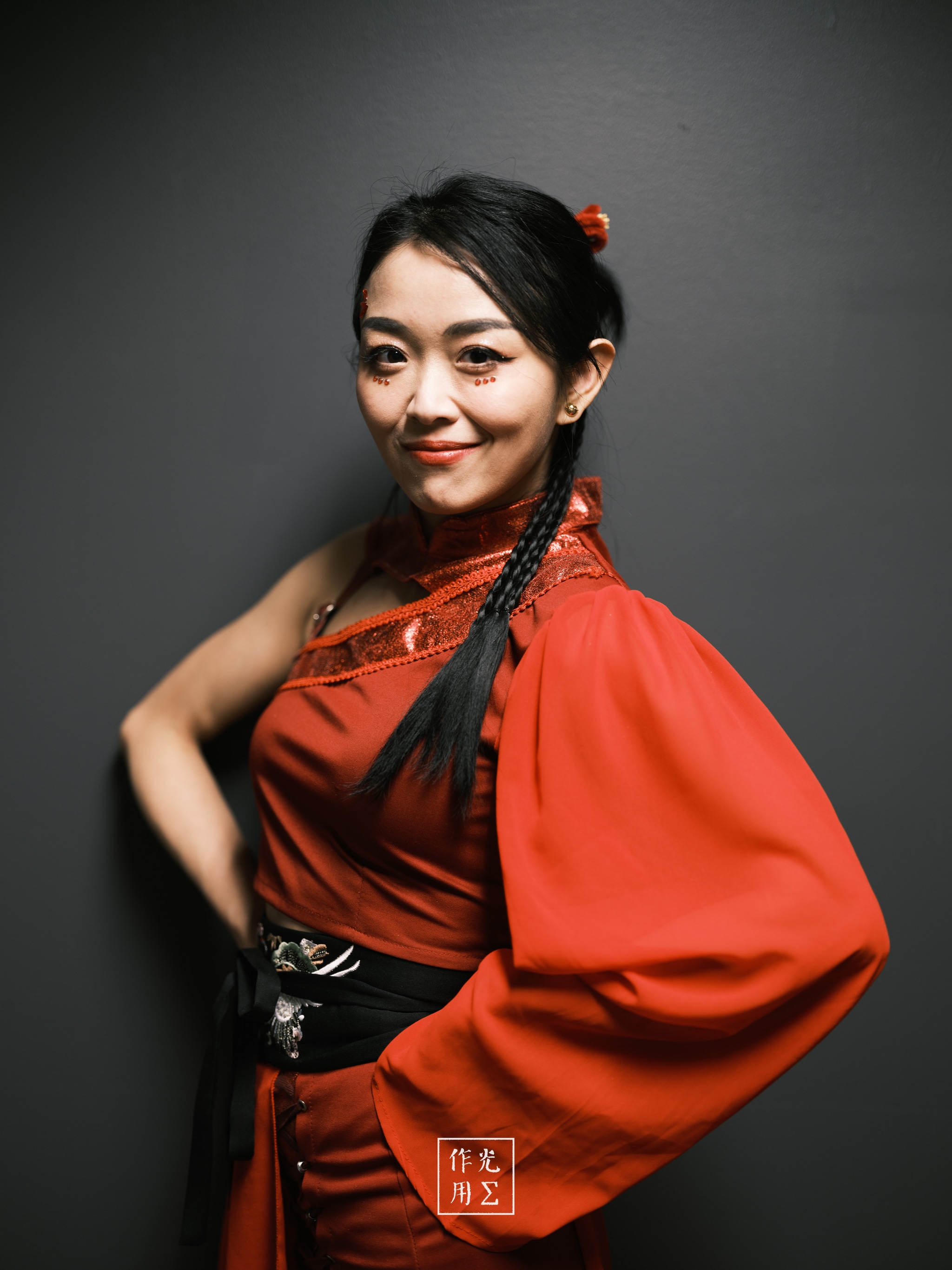 Radiating confidence, she stands against the charcoal backdrop draped in a flowing scarlet robe that cinches at the waist with ornate black embroidery. Twin braids cascade over her shoulder, each strand gleaming under the soft, directional light that sculpts her features and casts subtle shadows along her high cheekbones. A delicate crimson hairpin peeks from her dark silk bun, echoing the robe’s rich hue. Her knowing smile and poised hand on hip convey quiet strength and timeless elegance, turning the simple studio setting into a stage for striking, lacquered beauty