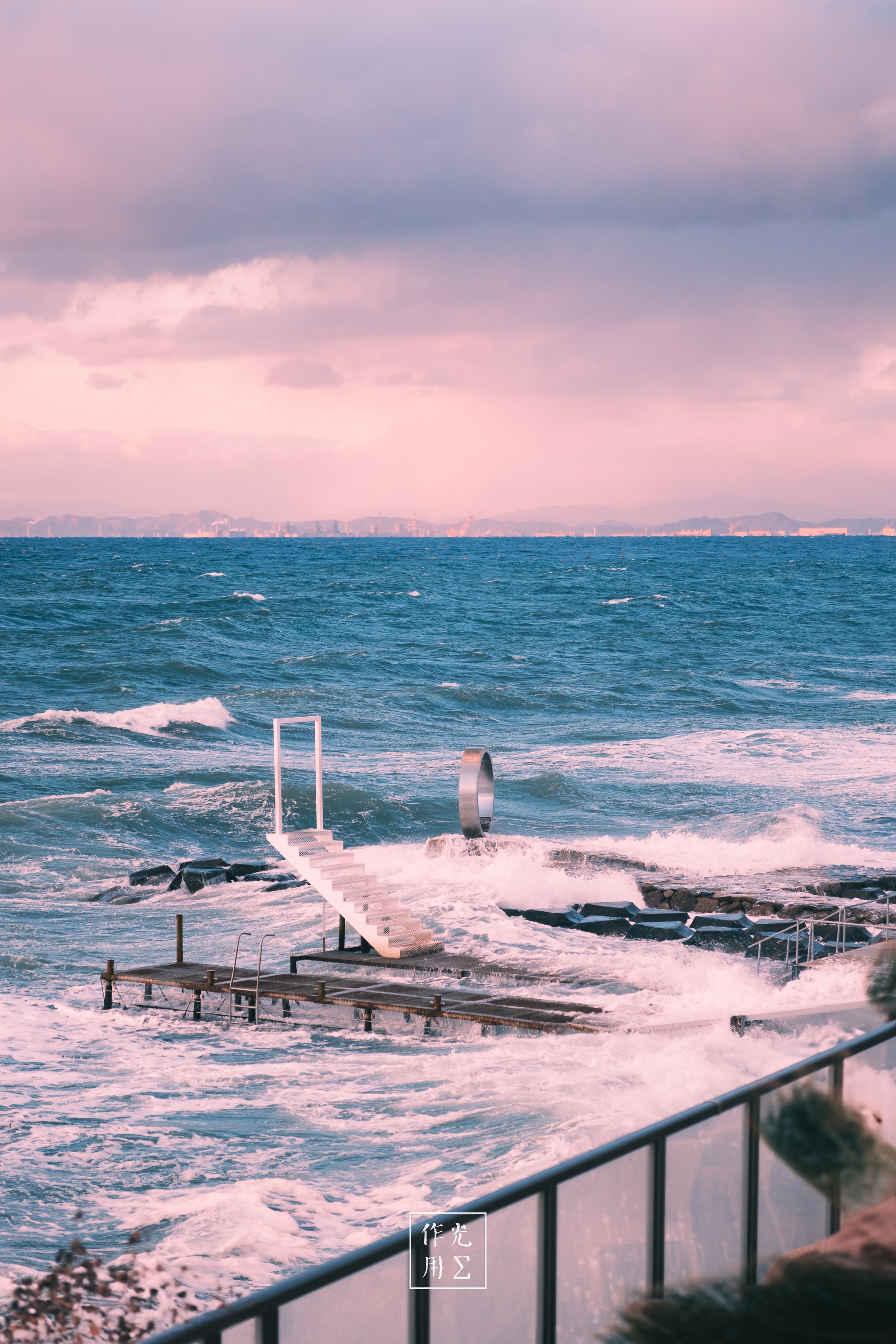 Surging foam lashes a weathered pier crowned by a lone diving ladder, its white railings streaked with salt. A restless cobalt sea churns beneath a blush-pink sky, clouds swirling in lavender and rose. Splintered planks dip beneath each wave’s crest while a wind-bent railing in the foreground frames the untamed dance of water and wood. Distant hills melt into the horizon’s rosy haze, lending a dreamlike cast to the raw, tempest-tossed energy