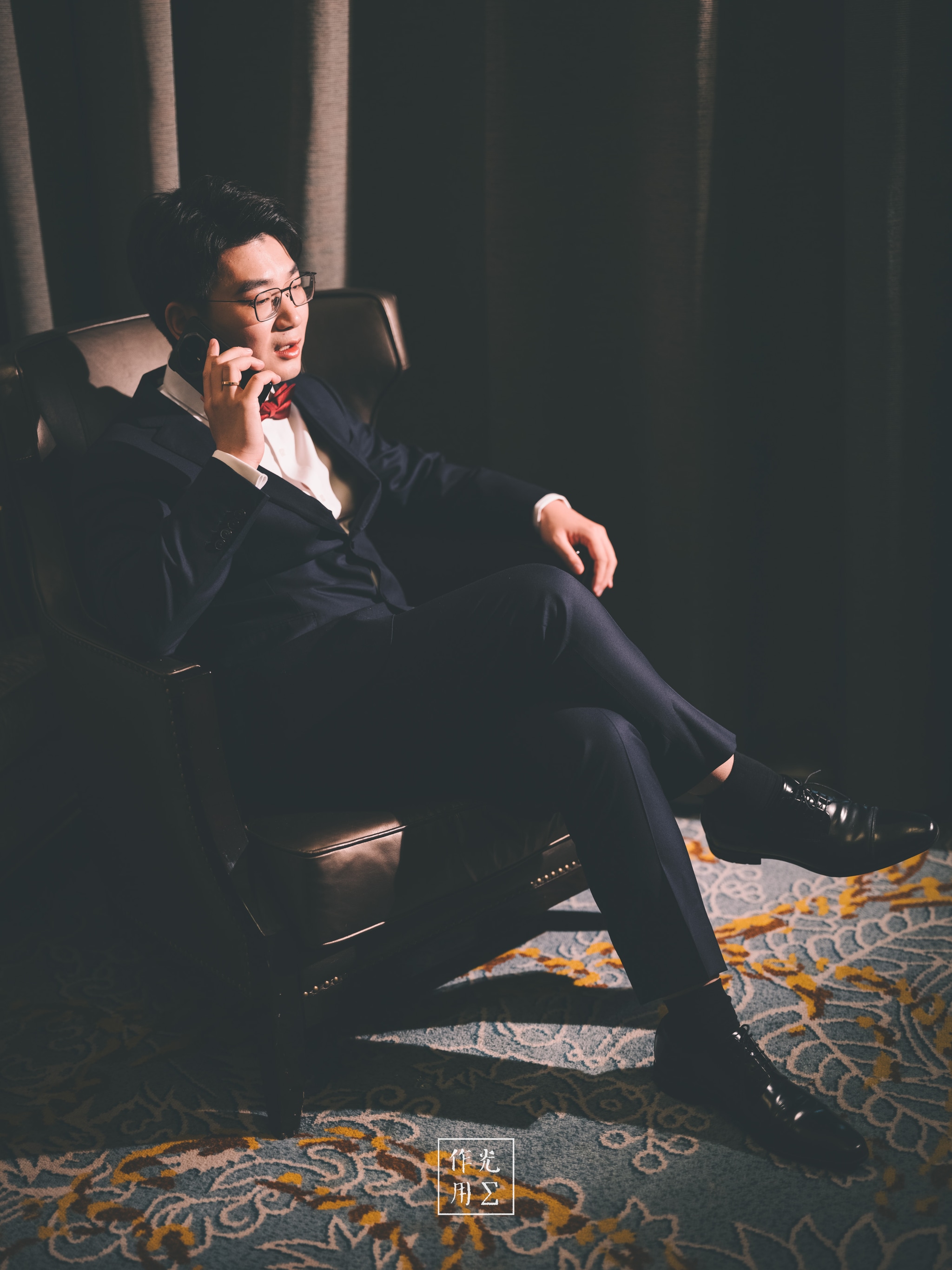 Leaning back in a deep leather armchair, he clasps a slim smartphone to his ear, the glow of its screen tracing the sharp line of his jaw His midnight-blue suit catches the soft lamplight, each fold and seam rendered in rich shadow and subtle sheen Behind him, heavy velvet drapes fall in brooding, cathedral-like folds, absorbing warmth and creating a cocoon of hushed secrecy Below, an ornate carpet patterned with gold and slate-blue flourishes cushions his polished Oxfords, whose gleam hints at fastidious care A faint haze drifts through the air, lending the scene a film-noir tension—as though every whispered word might unravel a high-stakes deal