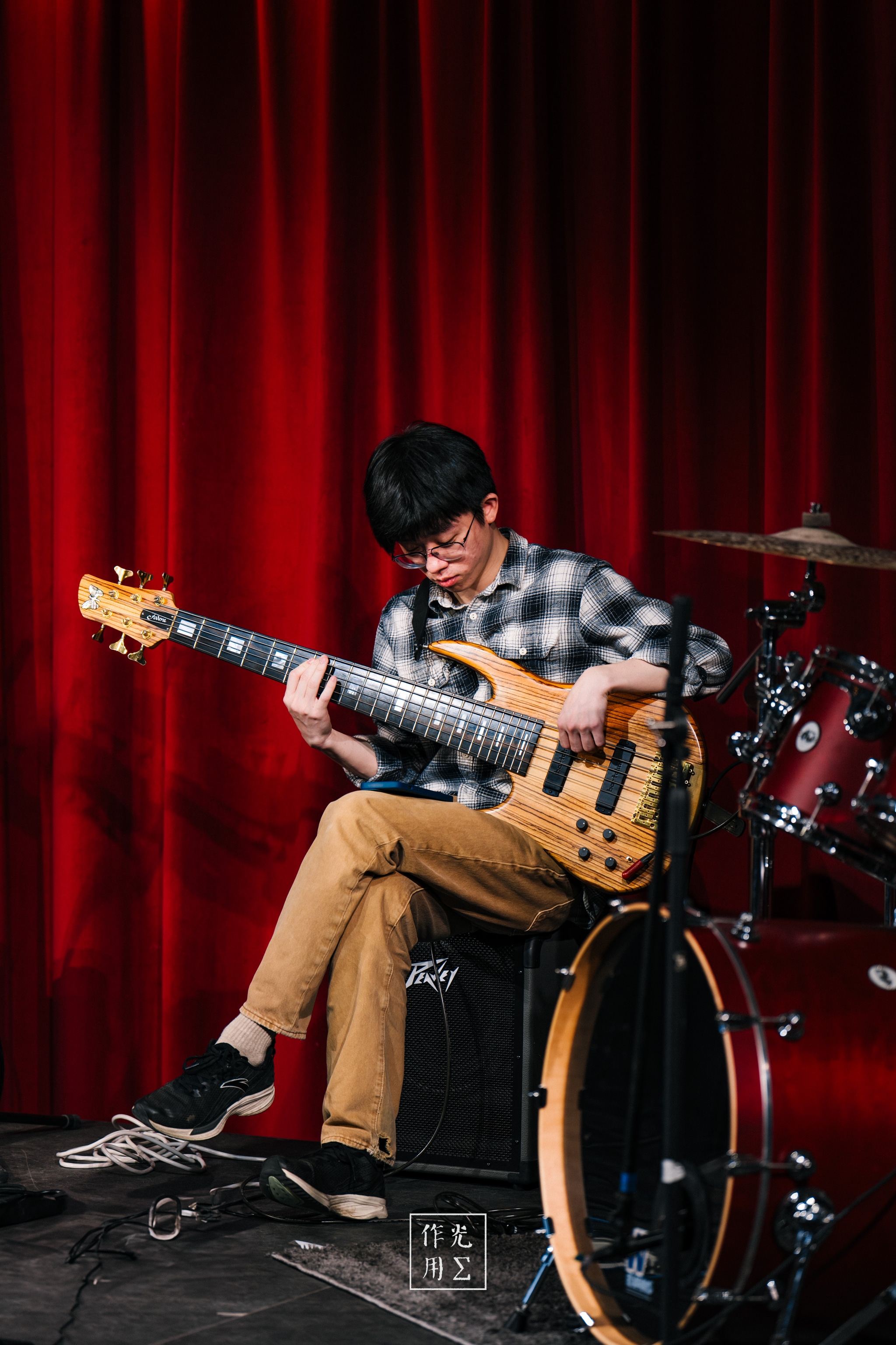 Bathed in the glow of muted amber spotlights, the bassist leans into each note, fingers dancing along the honey-hued neck of his five-string. A glossy red velvet curtain drapes the background like theatrical blood, amplifying every wood-grain ripple on the bass’s body. His checkered shirt and khaki trousers offer a homespun contrast to the stage’s polished sheen, while silent cymbals and toms loom behind him, hinting at rhythms yet to arrive. The air hums with anticipation—intimate, raw, and ready to erupt