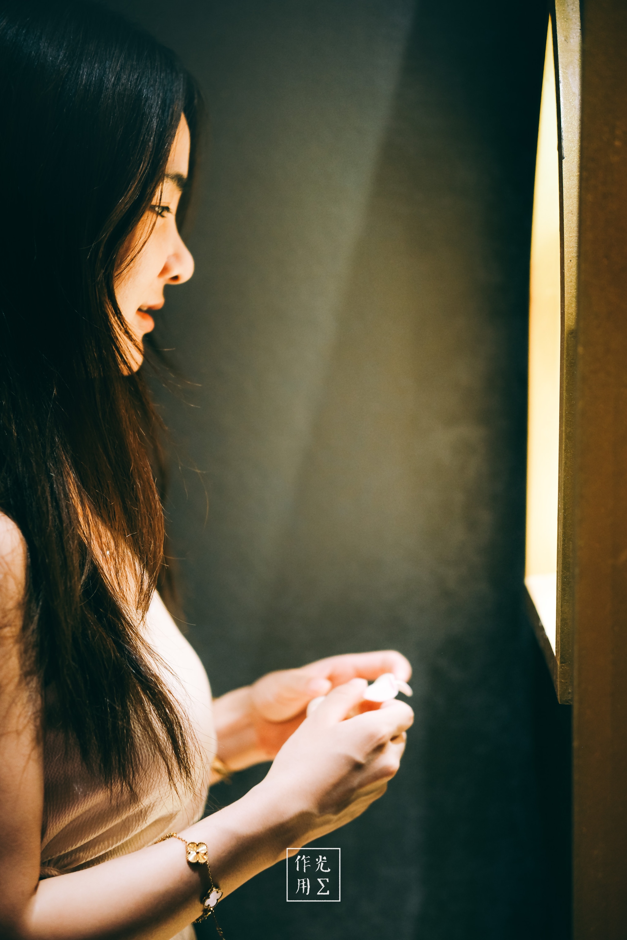 Bathed in amber glow, her profile emerges with haunting clarity against the midnight-black wall Soft strands of hair cascade over porcelain skin, catching flickers of warmth as she leans closer to the illuminated panel Fingers poised in gentle anticipation, she seems suspended between curiosity and calm reverie The stark contrast between light and shadow sharpens her silhouette, turning a simple glance into a moment of intimate wonder