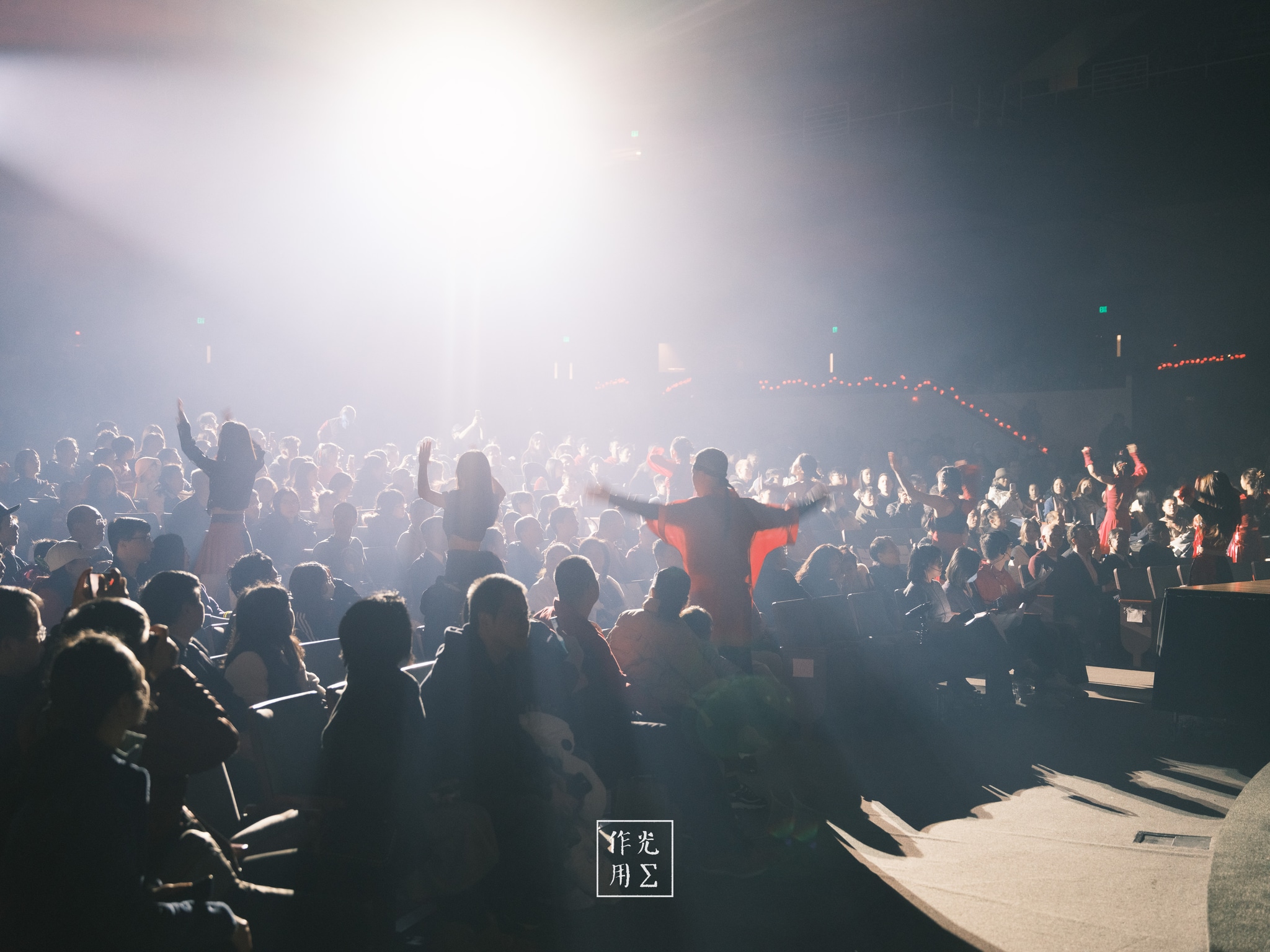 Jets of white light pierce through a haze of swirling dust, casting the crowd into sculpted silhouettes. A lone figure in a crimson shirt stands center stage, arms outstretched as if conducting the electric energy rippling through the arena. Faces blur into shadows and glimmering flecks in the air, each beam carving out momentary halos above waving hands. The atmosphere crackles with anticipation—raw, kinetic, and drenched in the promise of an unforgettable crescendo