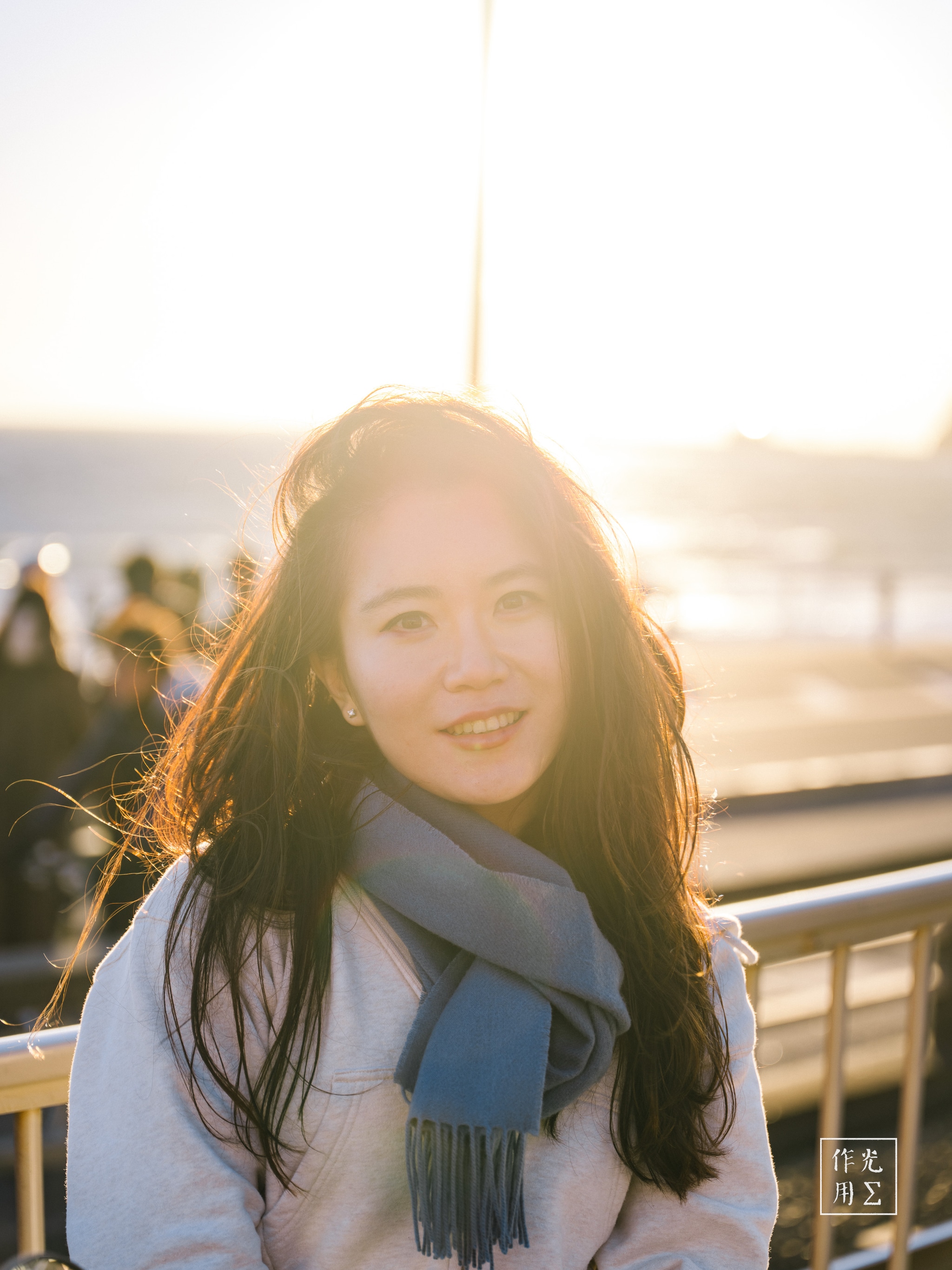 Basking in a halo of molten light, her dark tresses catch the dying sun and glow like spun amber. A soft breeze teases her scarf, its muted blue hue a cool counterpoint to the warm, honeyed tones of the sky and ocean behind. Silhouetted figures drift along the railing in gentle focus, while a single beam of sunlight splits the frame, lending the scene an almost ethereal edge—half dream, half memory