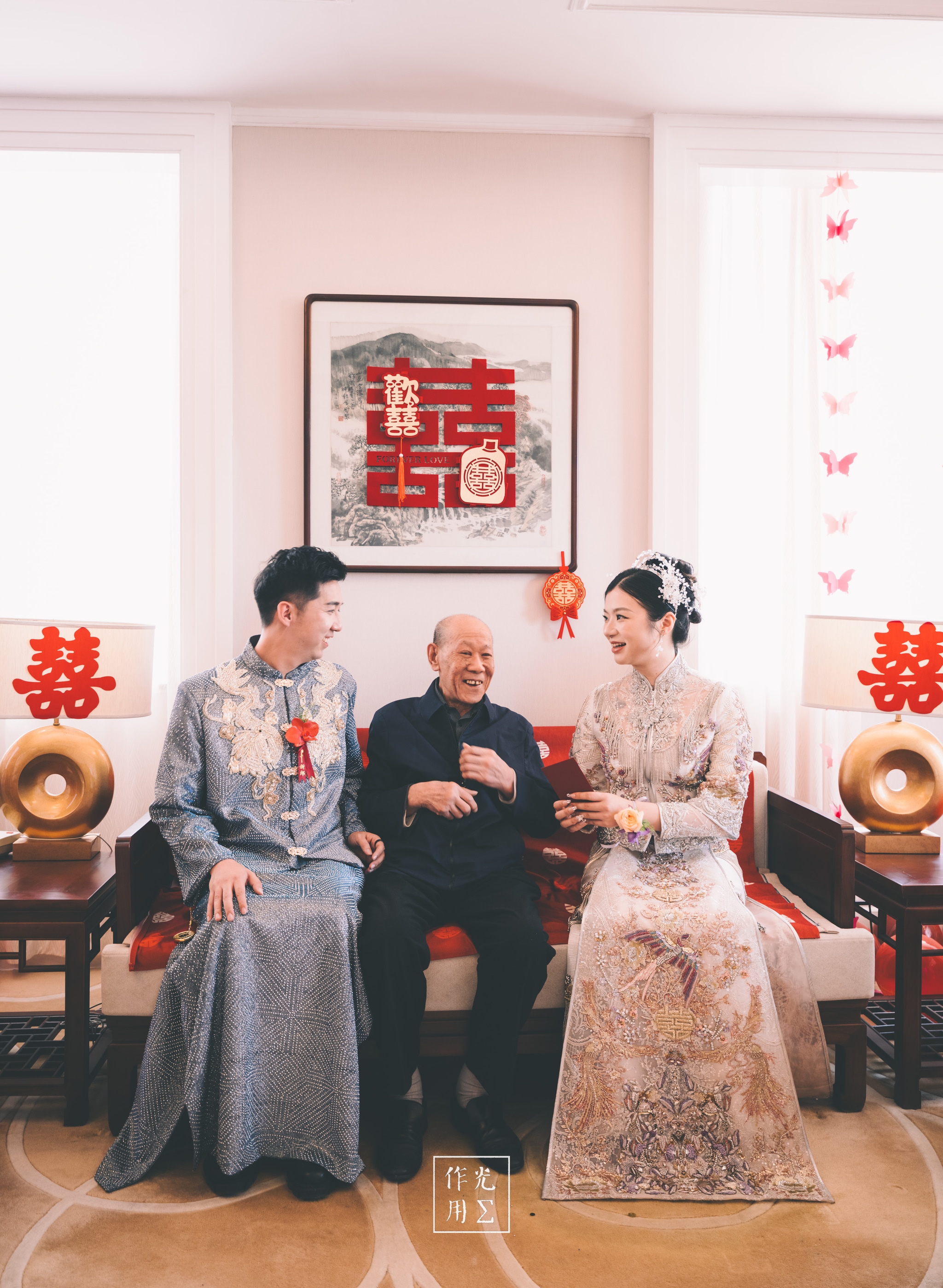 Silvery threads on the groom’s changshan catch the morning light while the bride’s ivory-embroidered gown shimmers beneath her delicate pearl veil. Between them, their grandfather’s warm smile creases into his cheeks as he lounges on the sofa, his dark jacket a grounding contrast to the pastel-pink walls. Flanking the trio, golden gramophone horns and scarlet “double-happiness” characters anchor the room, while a curtain of paper butterflies filters sunlight into soft, celebratory rays. Generations converge in laughter and ornate detail, weaving tradition and warmth into every stitch