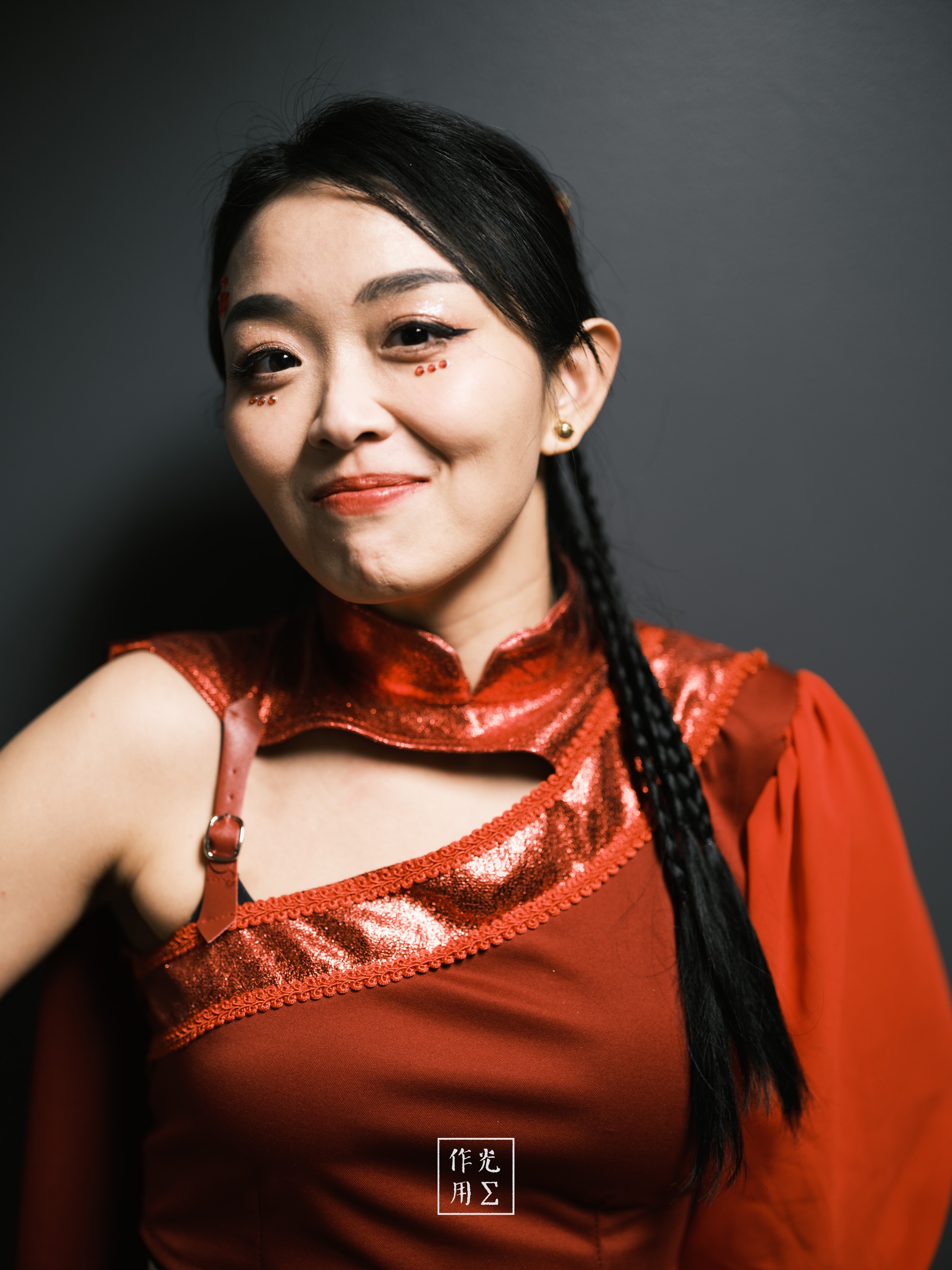 Radiating confidence under stark lighting, her asymmetrical crimson ensemble glints with metallic trim and carves a sharp silhouette against the charcoal backdrop. A single sleek braid drapes over one shoulder, framing a playful smirk and luminous eyes that dance with quiet mischief. A small calligraphic emblem at the hem adds an unexpected touch of contemporary elegance, anchoring the image’s bold, theatrical mood