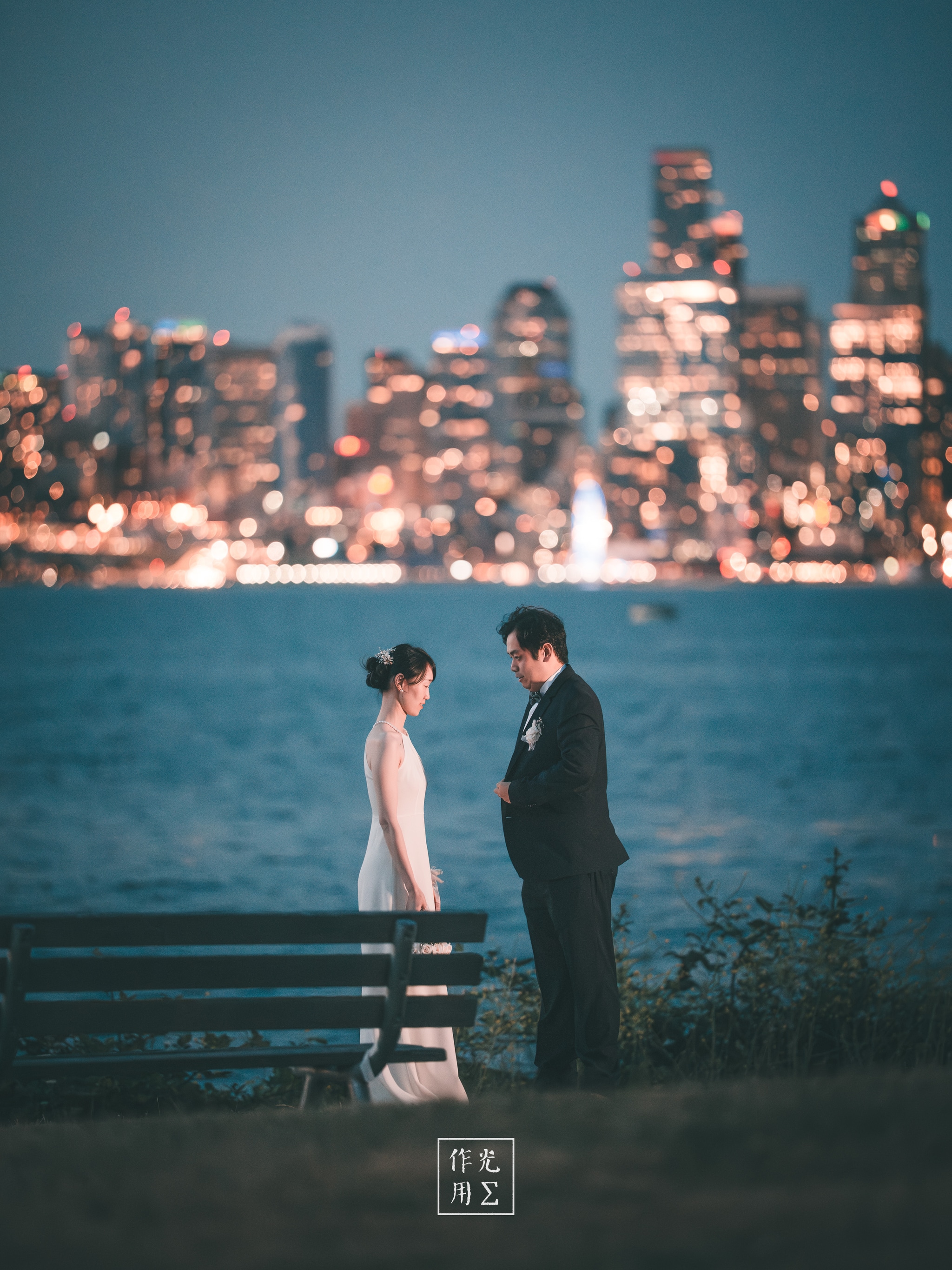 Glistening city lights ripple across the water as a couple stands at the water’s edge, the woman in a sleek ivory gown and the man in a tailored black suit A lone bench anchors the foreground, its empty slats echoing the quiet anticipation between them Behind, towering skyscrapers blur into a sea of gold and teal bokeh, turning the skyline into a dreamy, cinematic backdrop Cool night air drifts in off the bay, carrying hushed warmth of their whispered words and the faint hum of distant traffic Every glow and shadow frames their shared moment, balancing urban energy with intimate stillness