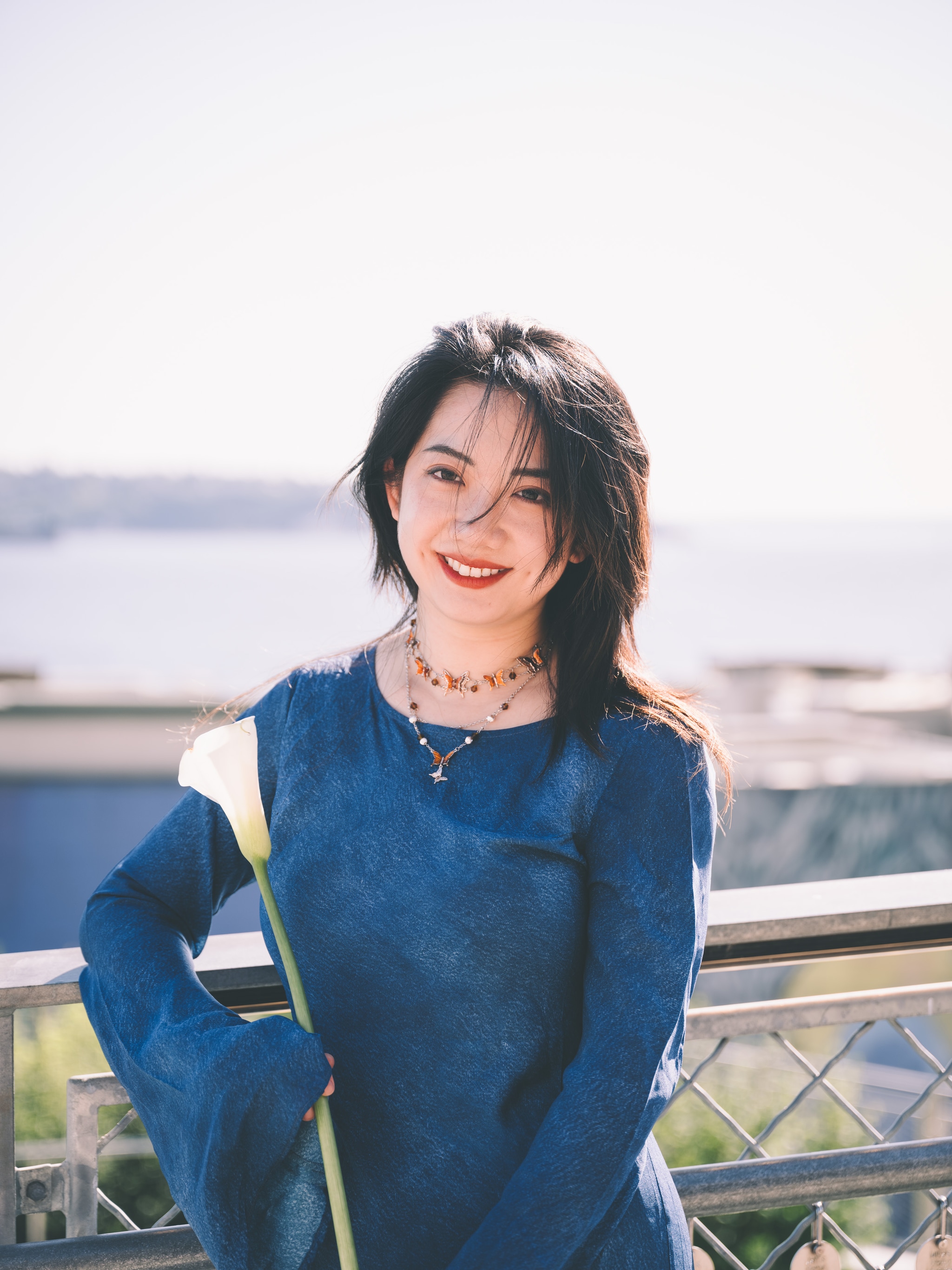 Breeze-tousled hair frames a radiant smile as sunlight glints on layered gold necklaces She leans against a railing overlooking a serene waterfront, her cobalt sweater’s soft texture contrasting with the metal lattice The hazy horizon and gentle ripples speak of tranquil afternoon air, amplifying her effortless poise and the scene’s quiet elegance