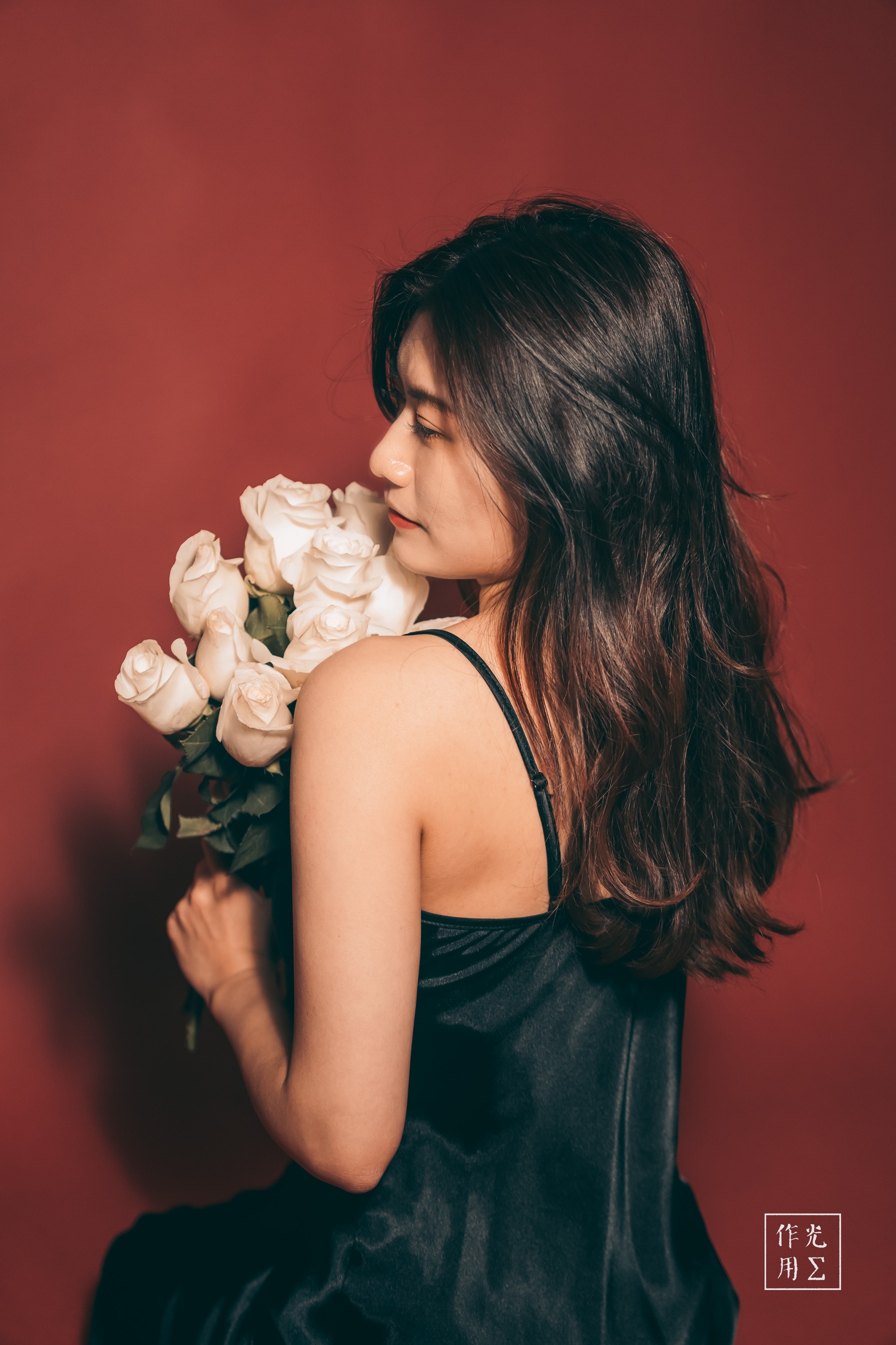 Crimson backdrop swirls warmth around her silhouette as she tilts her head toward a bouquet of pale roses. Silken black straps trace the curve of her shoulder, complementing the gentle fall of her dark waves. Petals almost glow against the rich red, while soft shadows lend an intimate hush to the scene. Her half-smile and closed eyes convey a quiet reverie, as if the scent of fresh blossoms has suspended time in a single, tender moment