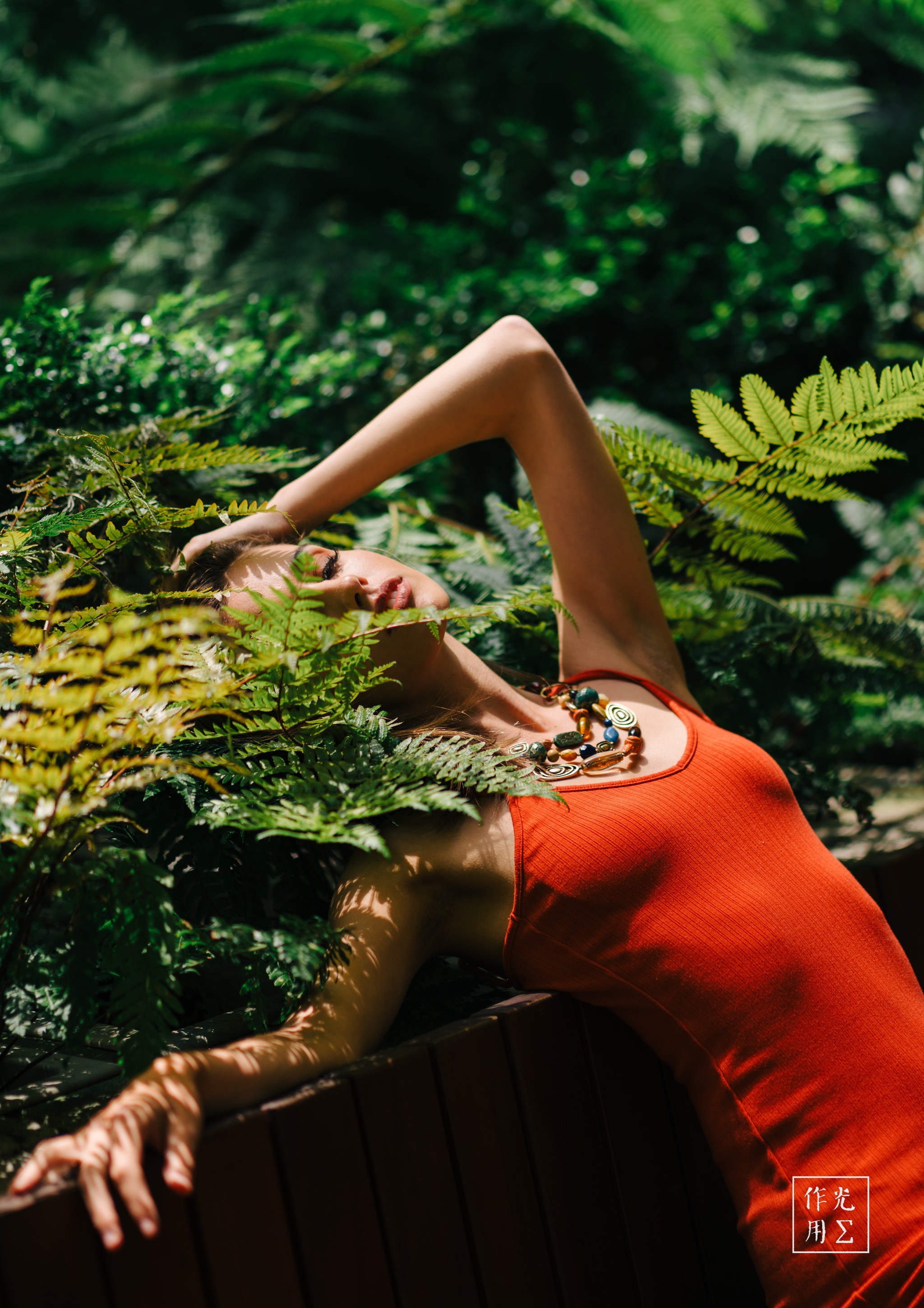 Basking in dappled sunlight, a woman draped in a vivid orange tank top reclines among emerald ferns. Her arm arches overhead, fingers brushing frond tips as filtered rays dance across her skin. A tangled necklace rests against her collarbone, echoing the forest’s organic curves. The lush greenery presses in, cocooning her in a hushed, dreamlike embrace where vibrant color and gentle shadows collide