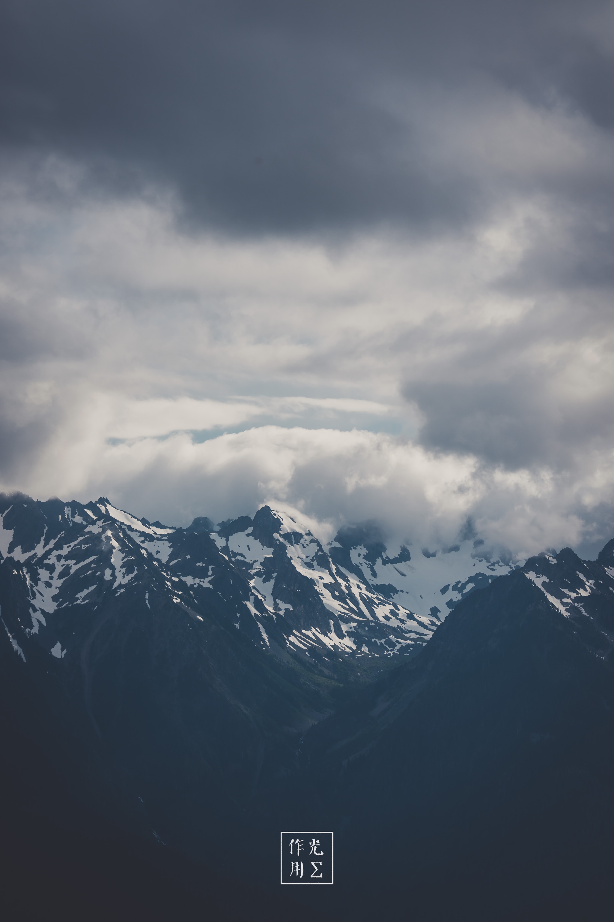 Jagged peaks thrust skyward, their snow-laced ridges cleaving through a churn of gunmetal clouds. A slender shaft of pale light seeps between storm-heavy layers, grazing icy summits and casting brooding shadows into the valley below. Wisps of mist coil around the mountains’ shoulders, heightening the raw, elemental drama of a landscape caught between light and looming tempest
