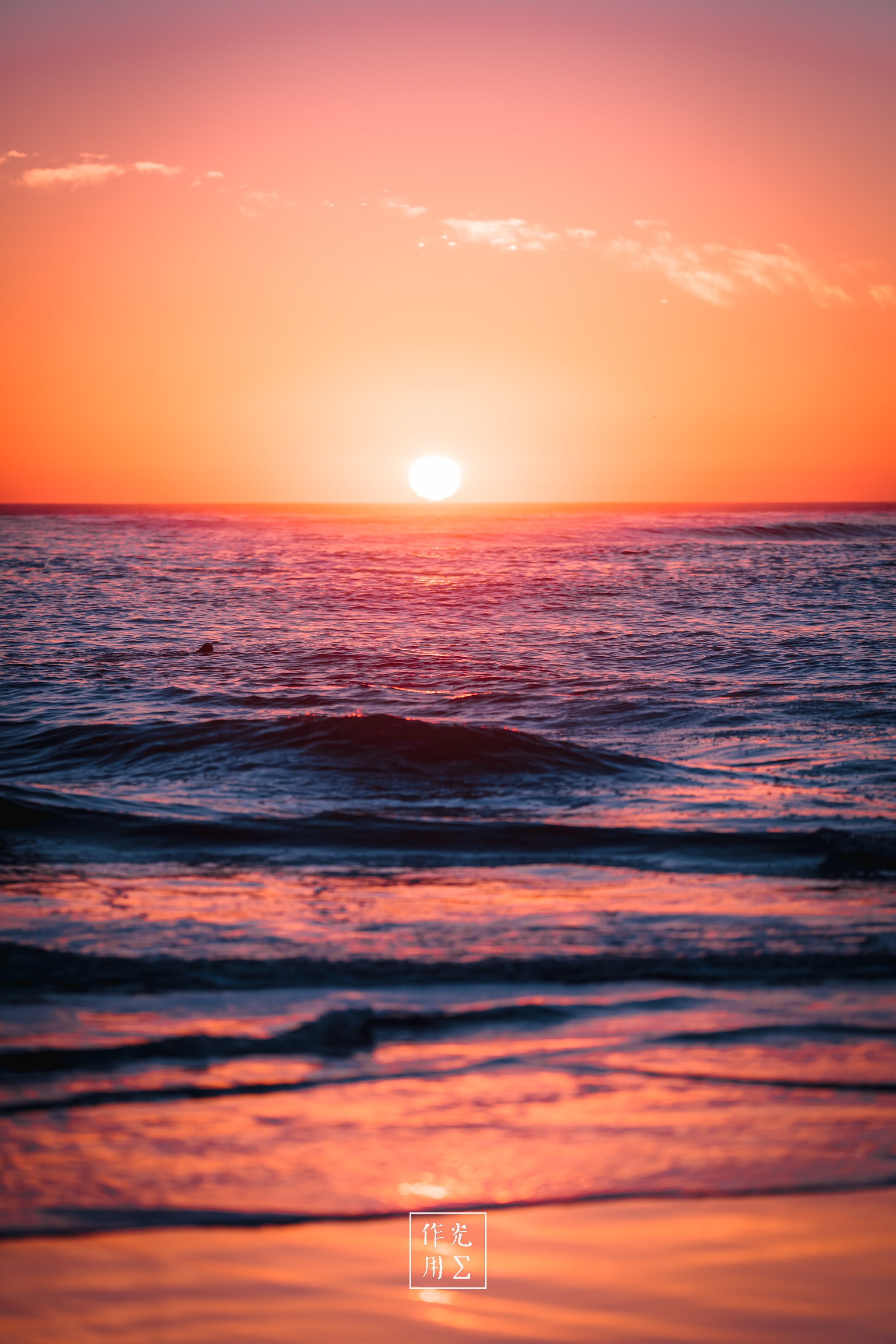 Sunlight spills like molten gold across a gently rippling sea, each wave tipped in rose and ember as the sun sinks toward the horizon. A soft haze of coral and lavender stretches overhead, blurring the boundary between sky and water. Wisps of cloud catch the last glow, their edges glowing neon against the deepening dusk. At the shoreline, translucent waves unfurl in rhythmic whispers, leaving wet sand shimmering with every hue of the fading day. A delicate artist’s seal in white punctuates the lower center, grounding this fleeting moment in timeless calm