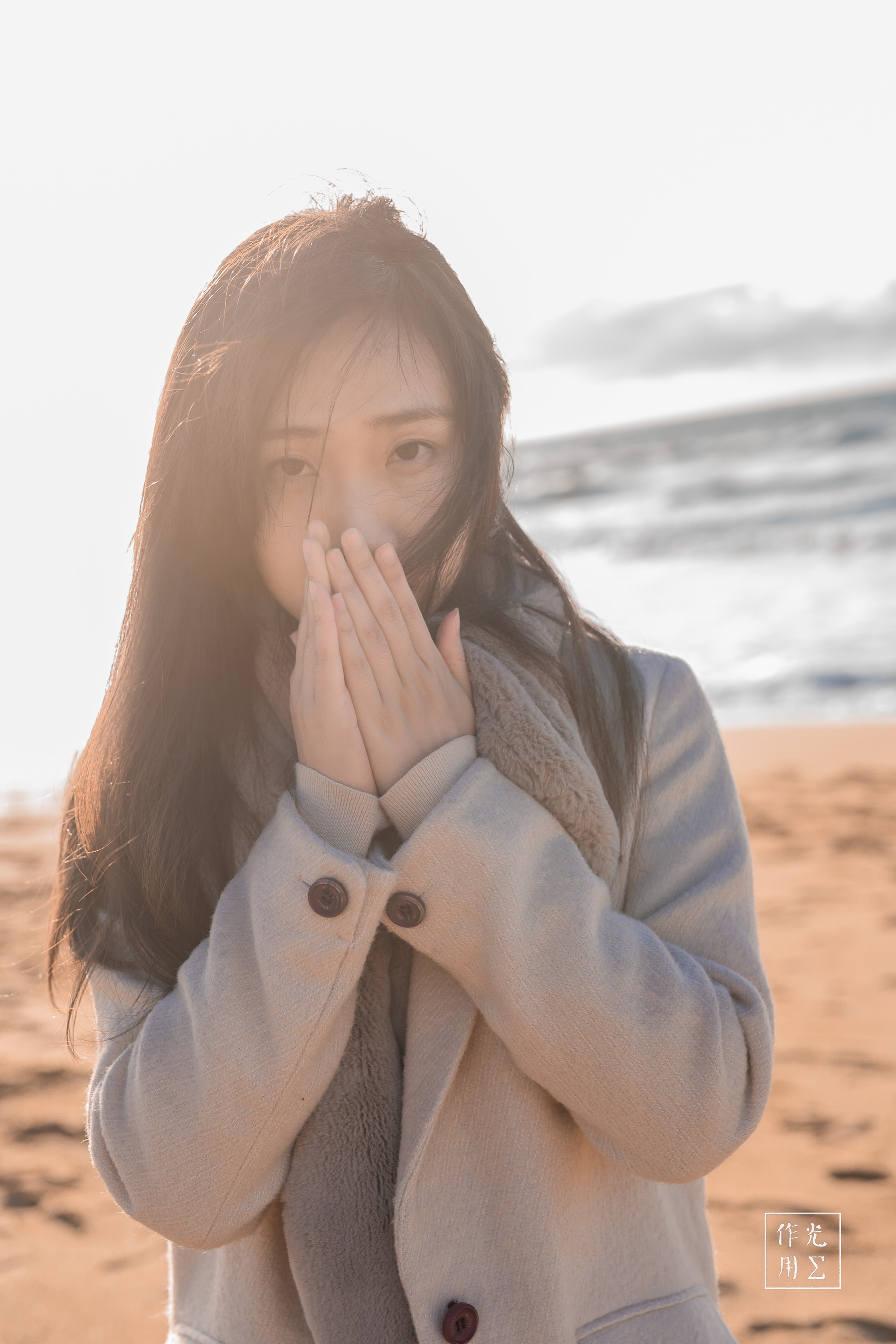 Bathed in a soft, golden-hour glow, she stands at the water’s edge in a tailored ivory coat, hands pressed to her lips against a brisk seaside breeze. Strands of dark hair drift across her face, catching flecks of sunlight like liquid amber. Behind her, gentle waves roll in under a pale sky, their muted turquoise and sandstone tones echoing the calm yet brisk mood. The quiet tension between her protective posture and the vast, open shoreline creates an intimate, almost secret moment—where warmth and chill, light and shadow, meet in a single, breath-caught frame