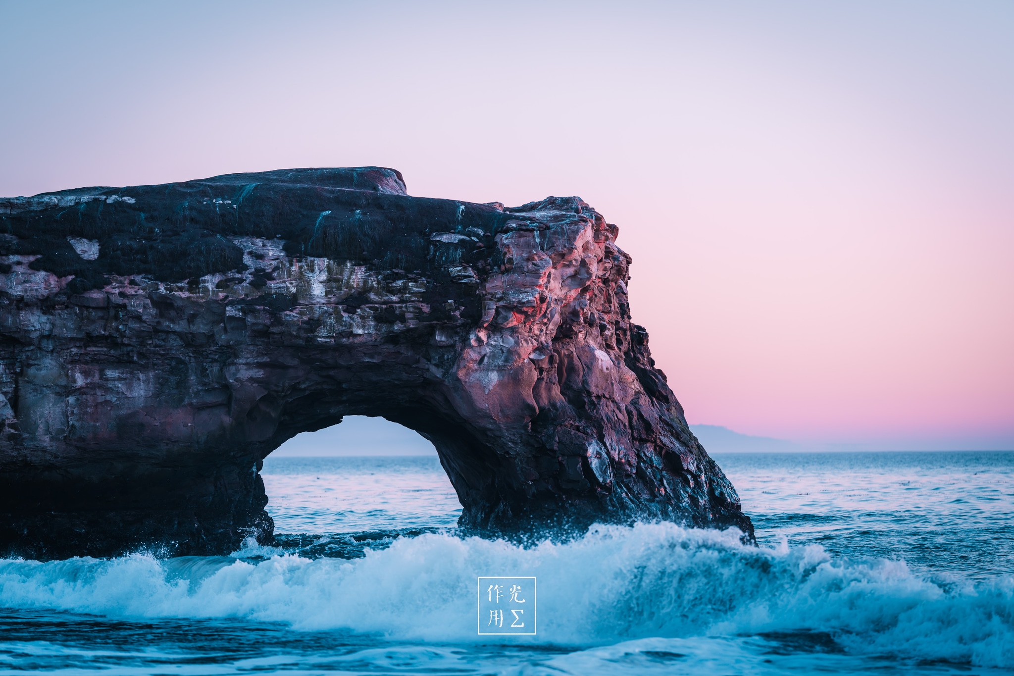 Beneath a pastel sky melting from lavender to blush, a rugged coastal arch juts into frothing teal surf. Golden-pink light bathes the limestone’s weathered contours, carving every crevice in warm relief. Rolling waves whisper through the opening, their foamy edges catching the sunset’s glow. The contrast of sharp stone against soft twilight sky lends the scene an otherworldly calm, as if time itself hesitates at this threshold between land, sea, and sky