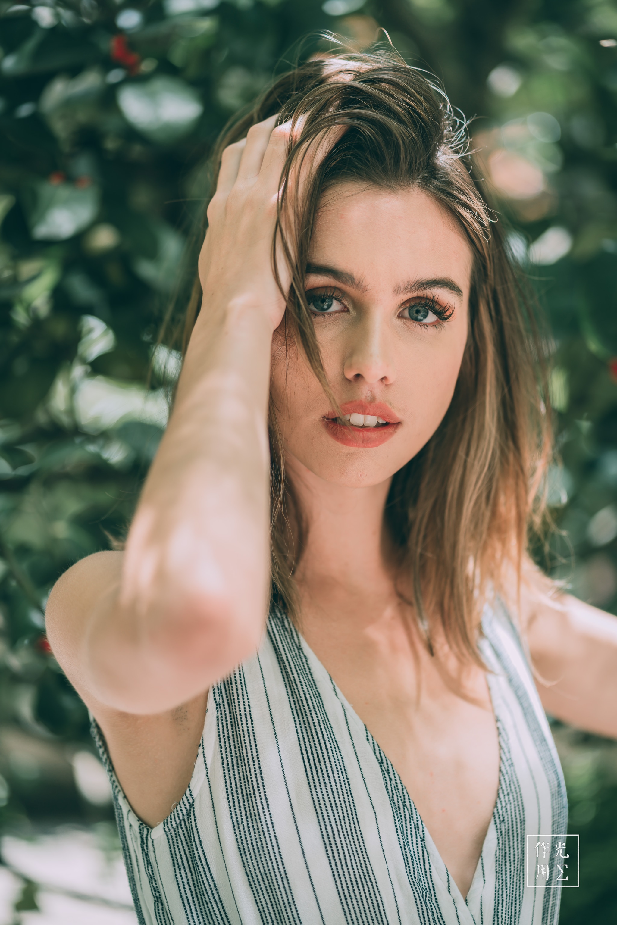 Sunlight filters through lush leaves, dappling her tousled chestnut hair and warm complexion. Slender fingers sweep strands aside, unveiling vivid green eyes that gleam with quiet confidence. Soft linen stripes in pale teal and ivory hug her shoulders, offering a breezy contrast to the deep foliage. The shallow depth of field blurs the surrounding greenery into a painterly haze, evoking the hush and mystery of a hidden garden