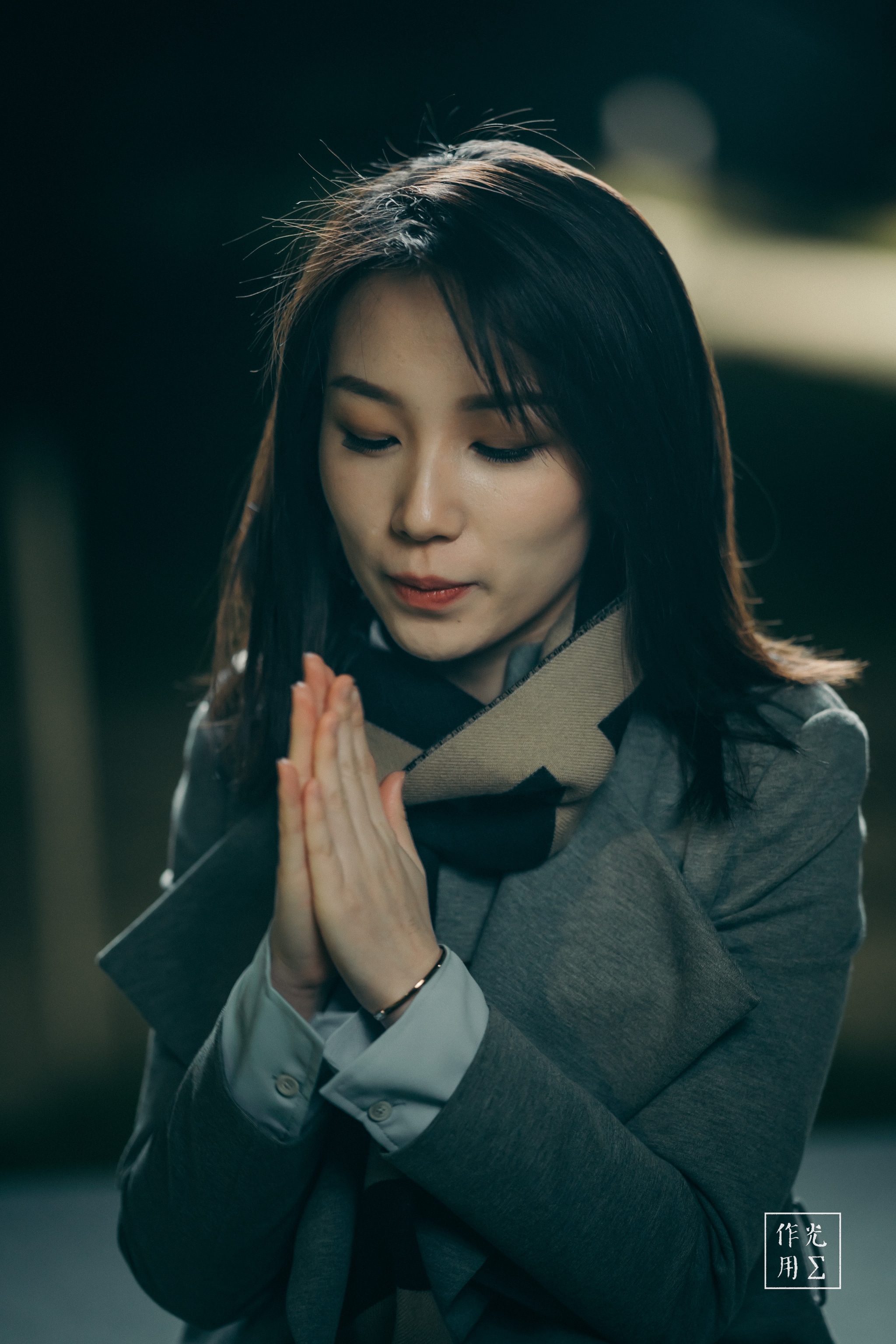 Warm lamplight spills across her face, casting gentle shadows that cradle her closed eyes and softly curved lips. Fingers pressed together glide upward, accentuating the quiet stillness in the bridge of her nose and the arch of her brows. A charcoal-gray coat and striped scarf wrap her in muted winter tones, while a blurred background of distant lights hints at a cold evening street. The contrast between the soft glow on her skin and the deep shadows around her creates an intimate moment of calm focus, as if time itself has paused around her silent prayer